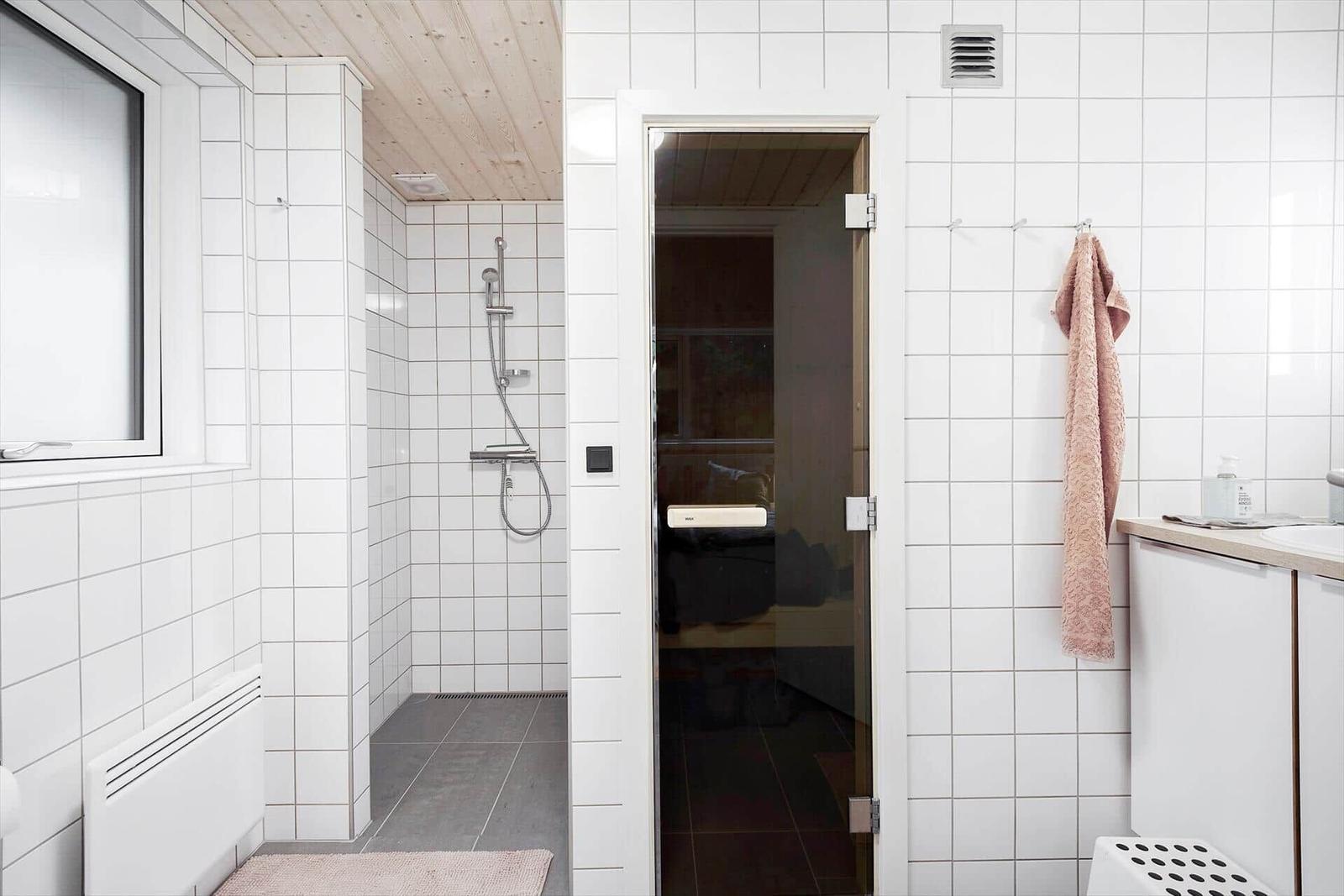 Bath with shower and sauna in background. White tiles and wooden ceiling.