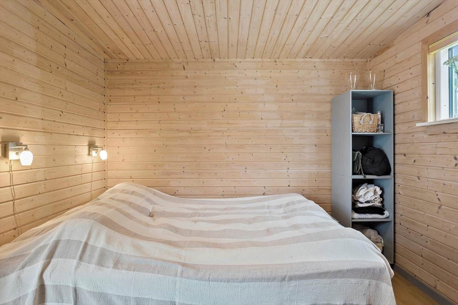 Schlafzimmer mit Holzwand, Bett und Regal. Fenster und Wandlampen sichtbar.
