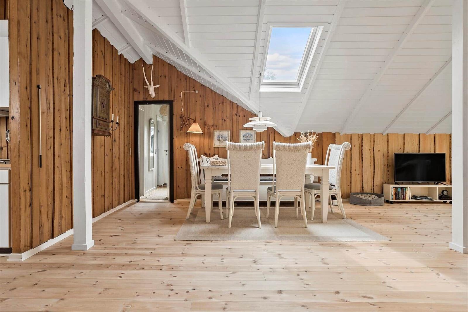 Dining area with white table and chairs under skylight. Wooden walls and floor. TV on stand.