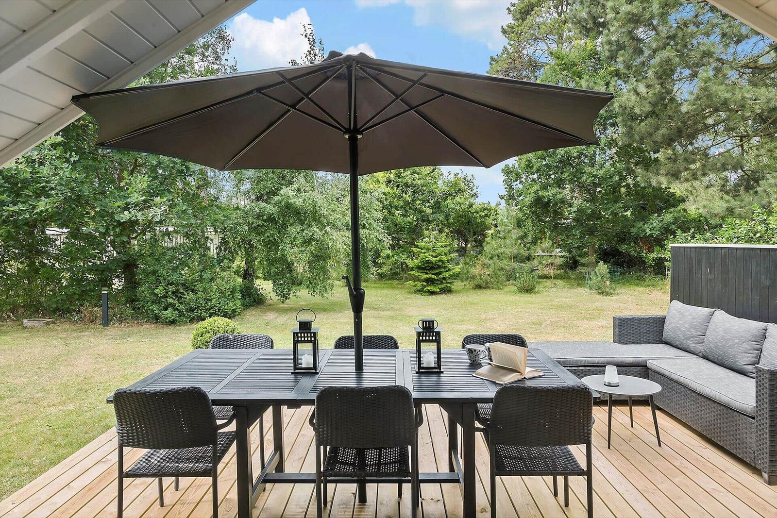 Terrasse mit Tisch, Stühlen und Sonnenschirm. Hintergrund: Grasfläche und Bäume.