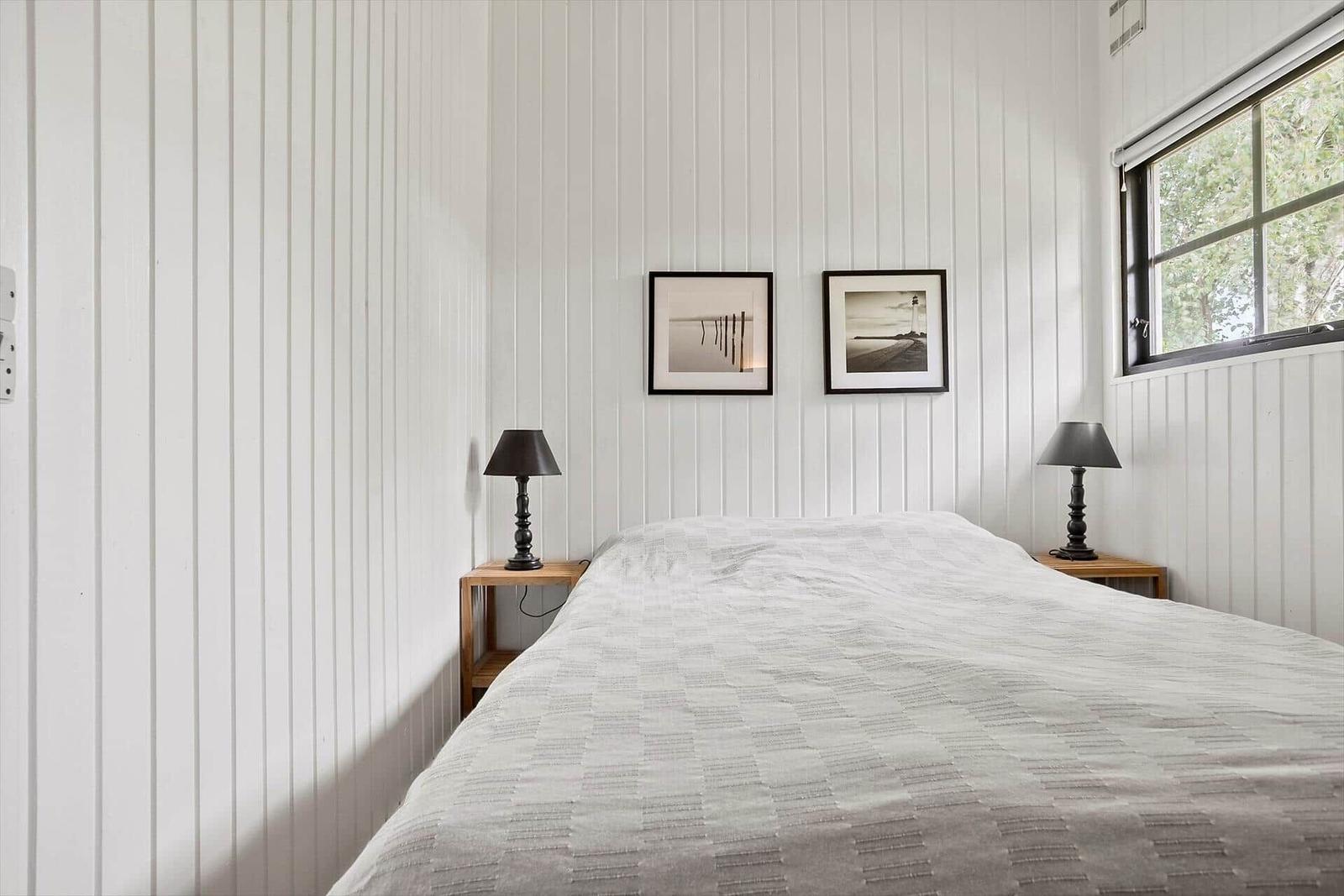 Schlafzimmer mit weiß verkleideten Wänden, Bett, zwei Nachttischen und Fenster.