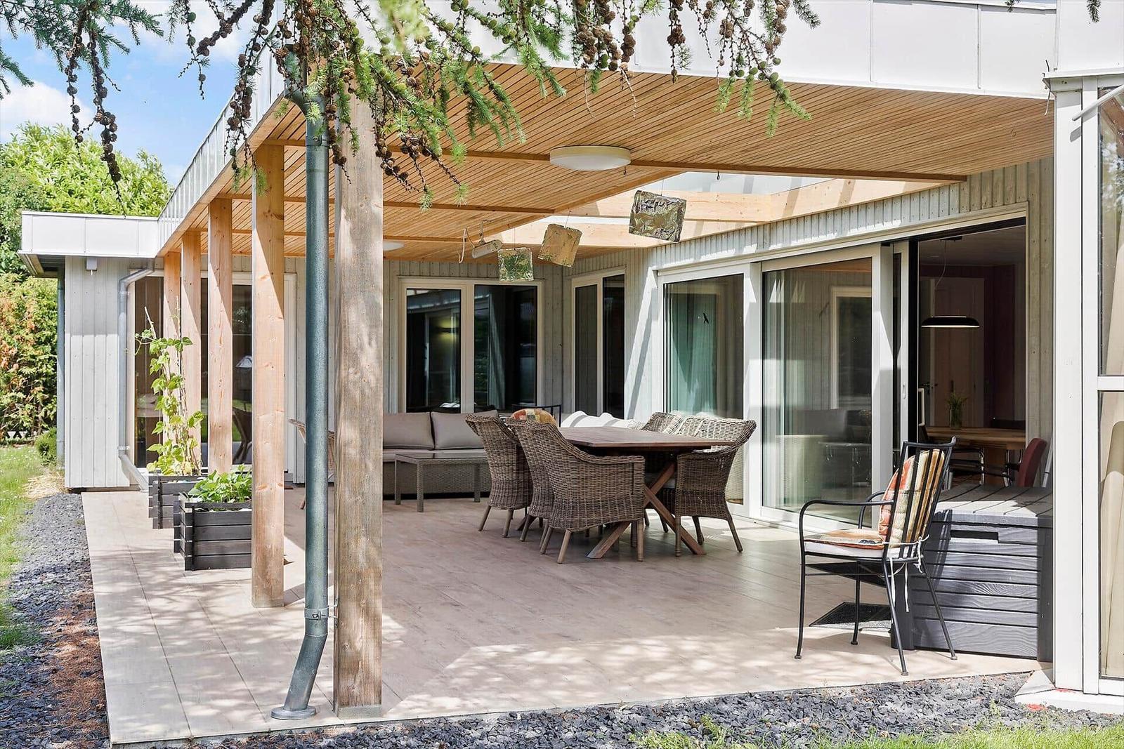 Terrasse mit Sitzgruppe und Tisch unter Holzüberdachung. Glasfront führt ins Haus.