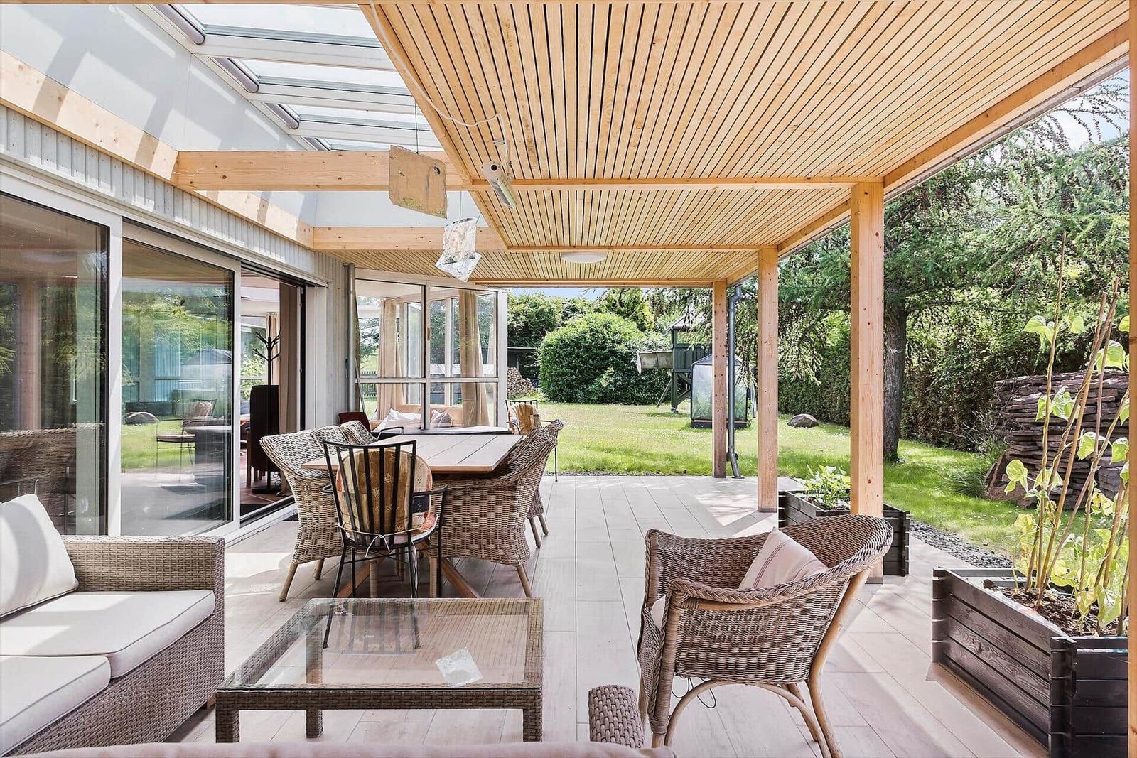 Terrasse mit Holzdecke, Sitzgruppe und Tisch. Grünfläche und Bäume im Hintergrund.