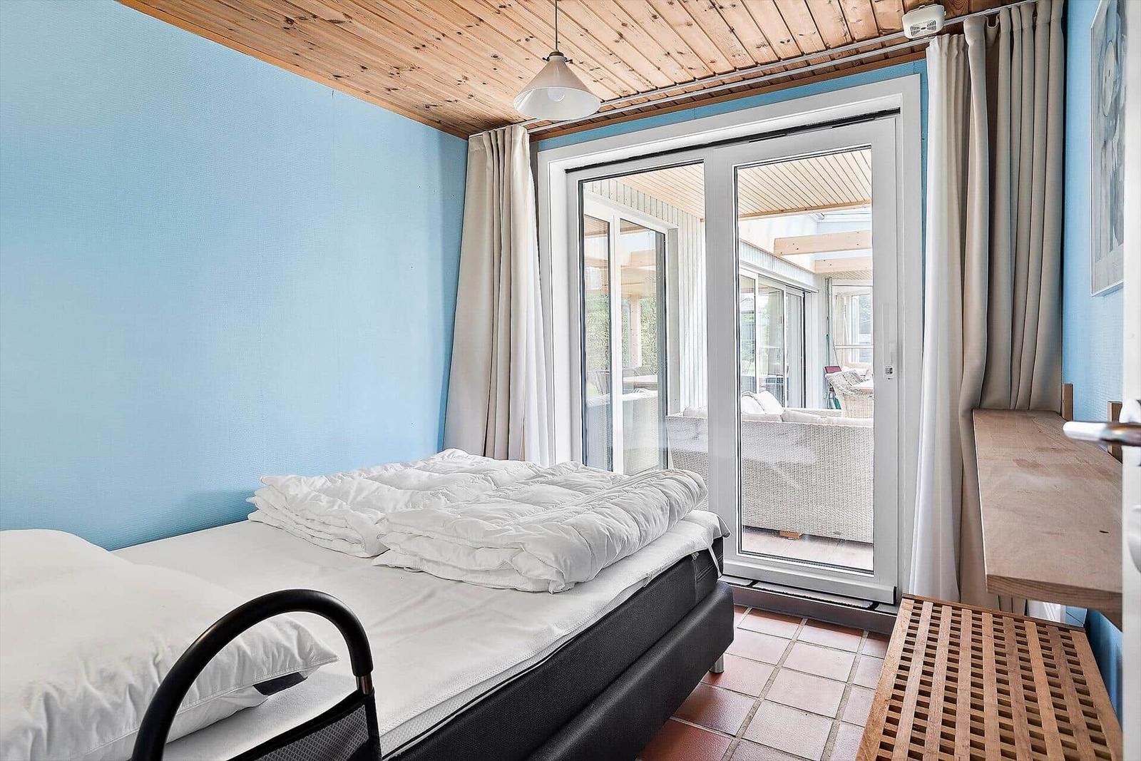 Schlafzimmer mit Bett, Glasfront und Terrasse. Holzdecke und blaue Wände.