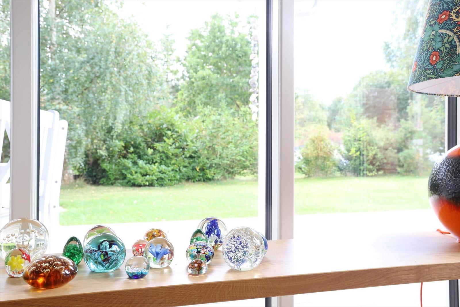Auf dem Fensterbrett stehen farbige Glasobjekte. Draußen ist ein grüner Garten sichtbar.