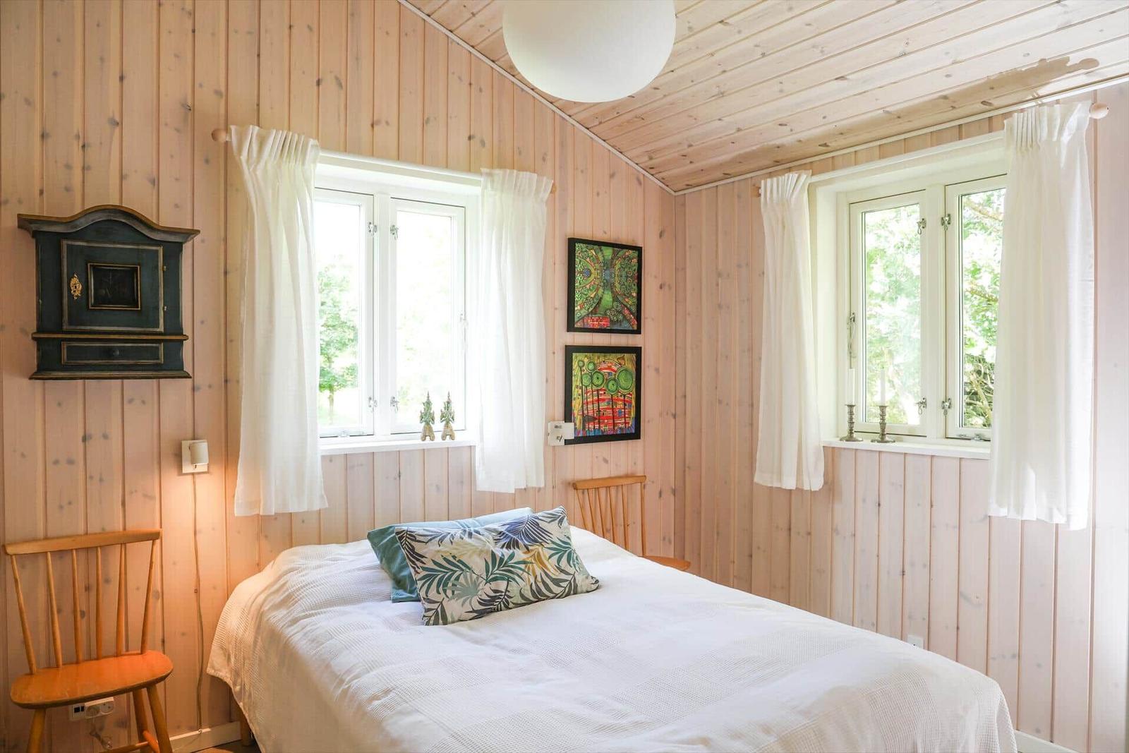 Schlafzimmer mit Holzwand, Bett, zwei Fenstern und Wandbildern.