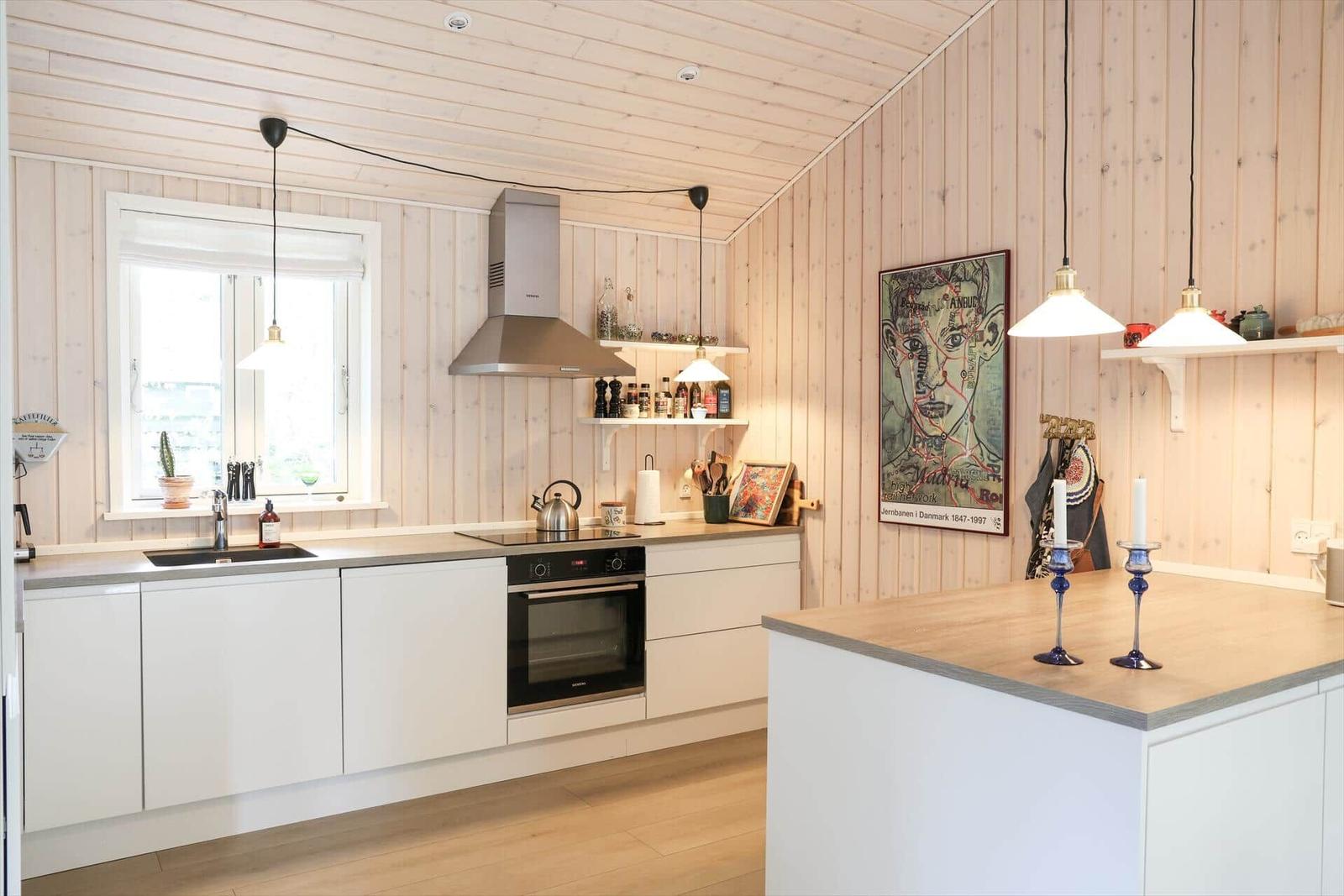 Küche mit weißen Schränken, Holzwand und Arbeitsplatte. Drei Hängelampen und Fenster.