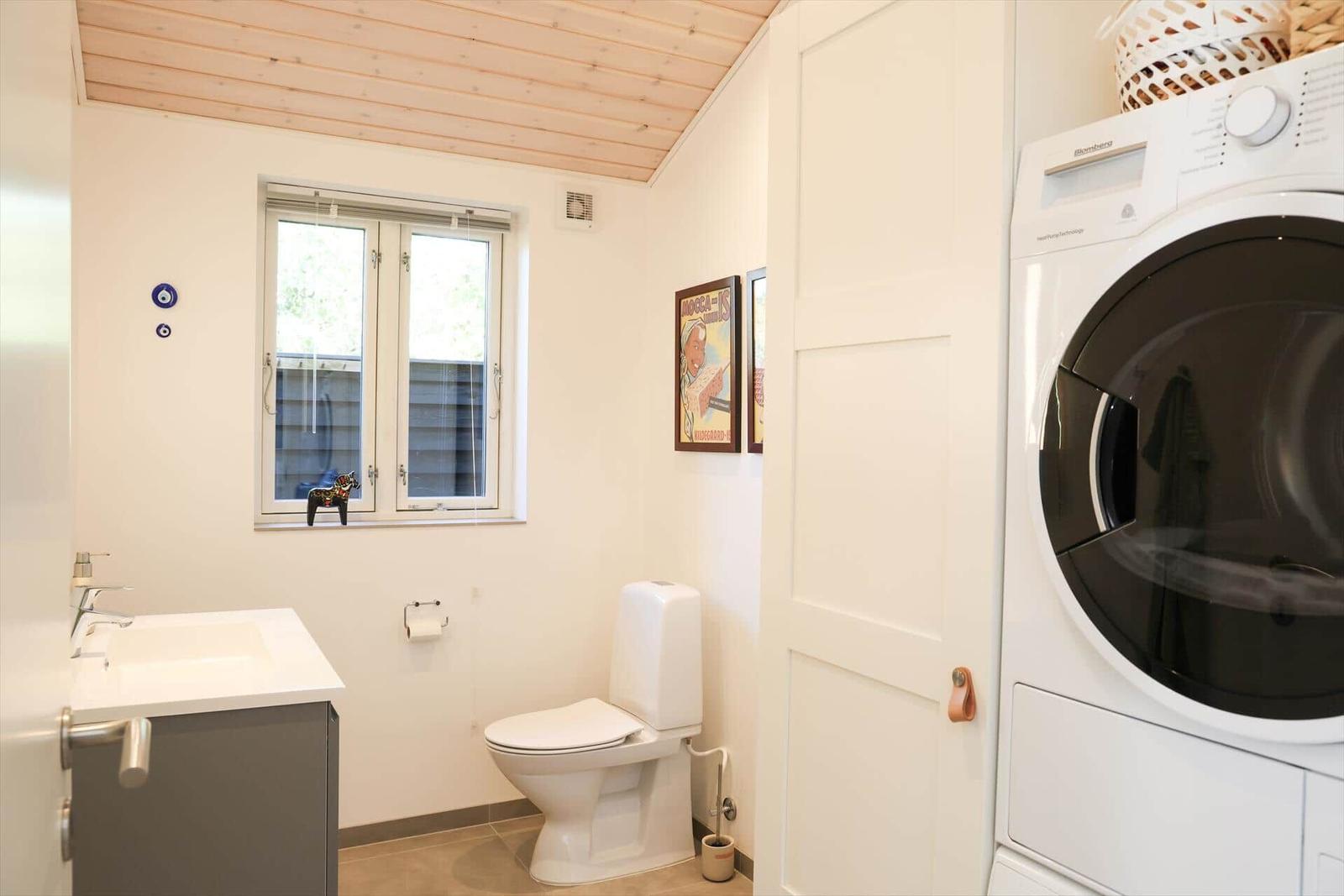 Badezimmer mit Toilette, Waschbecken und Waschmaschine. Holzdecke und Fenster mit Dekoration.