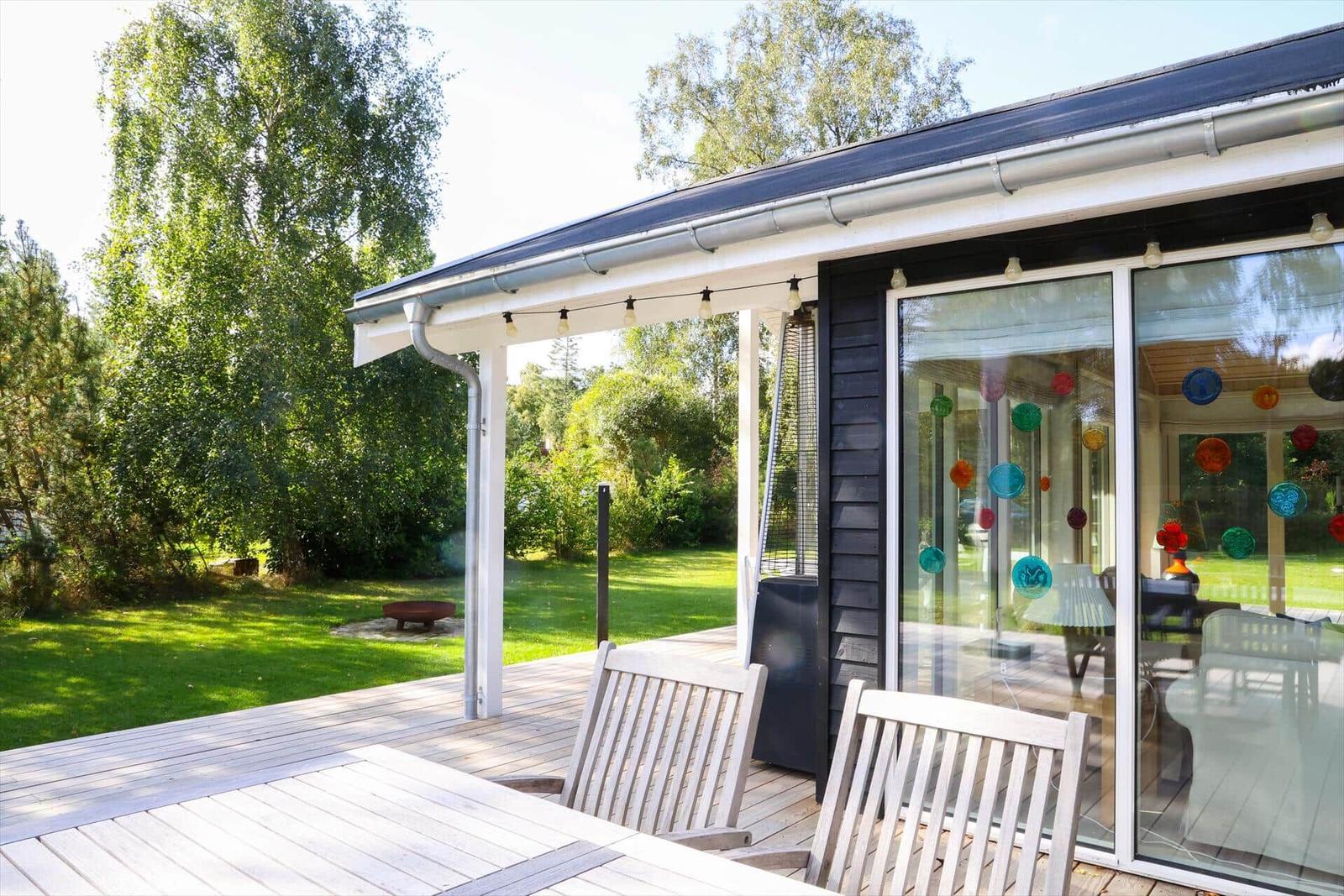 Terrasse mit Tisch und Stühlen, großes Fenster, Garten und Bäume im Hintergrund.