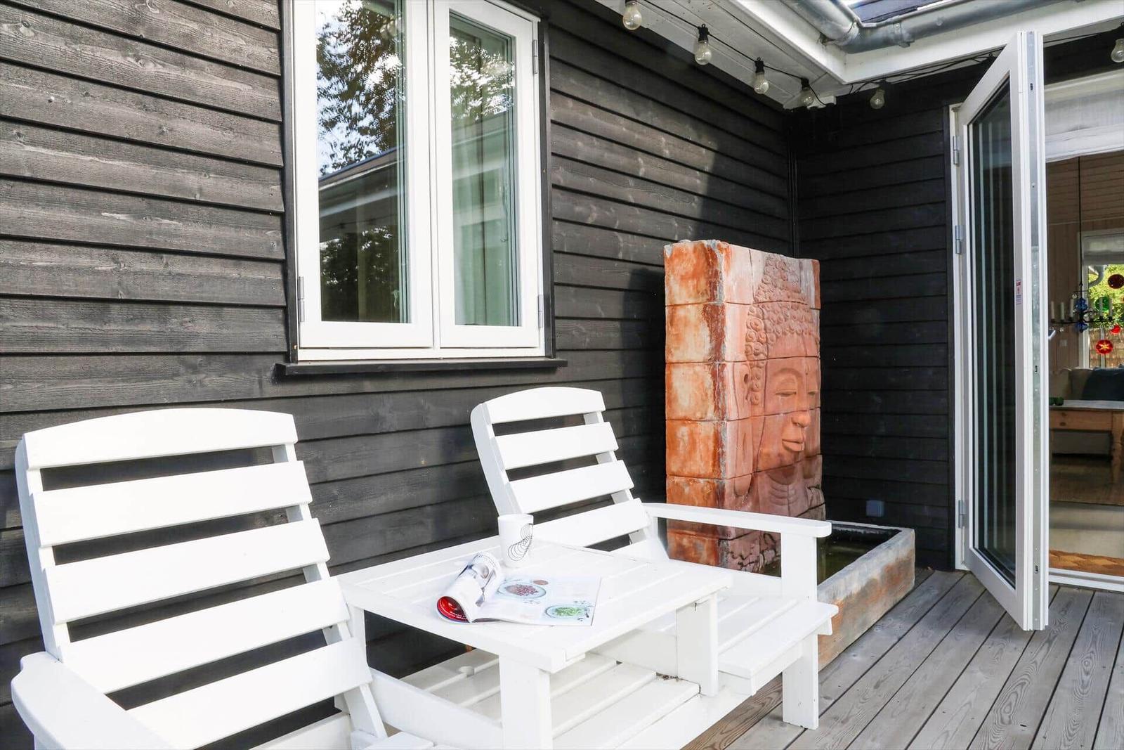Terrasse mit weißen Stühlen, Tisch und Buddha-Statue. Holzwand und Glastür sichtbar.