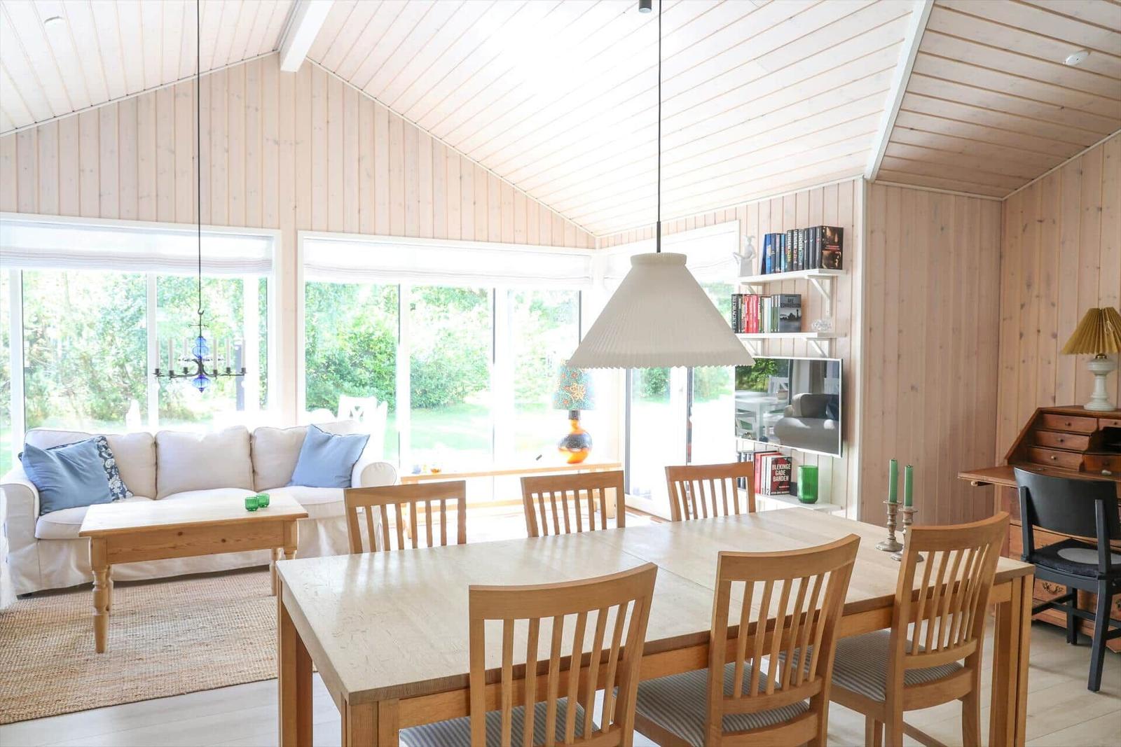 Ein Essbereich mit Holztisch und Stühlen. Im Hintergrund ein Wohnbereich mit Sofa und Fenstern.