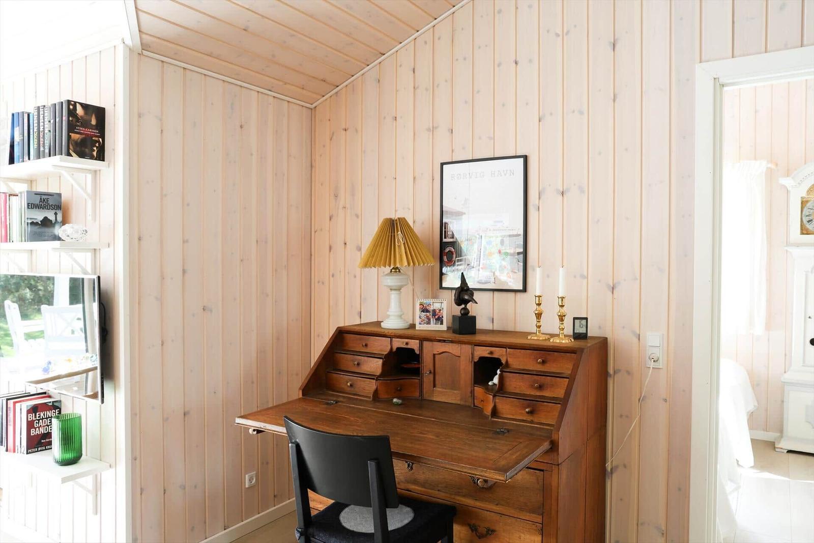 Schreibtisch mit Stuhl, Lampe und Bild an Holzwand. Bücherregal und Türöffnung sichtbar.