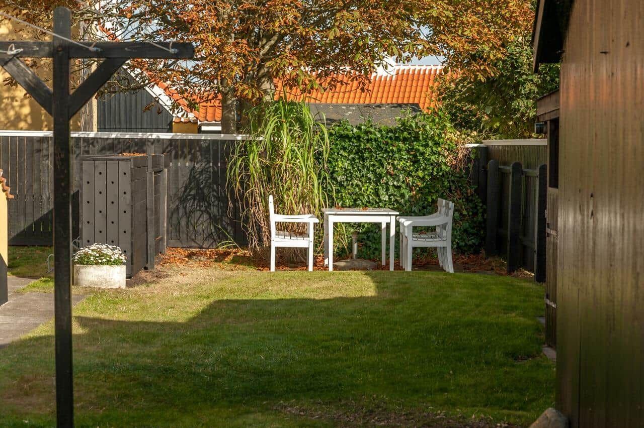 Hinterhof mit weißem Tisch und zwei Stühlen, umgeben von Gras, Bäumen und einer Holzumzäunung.