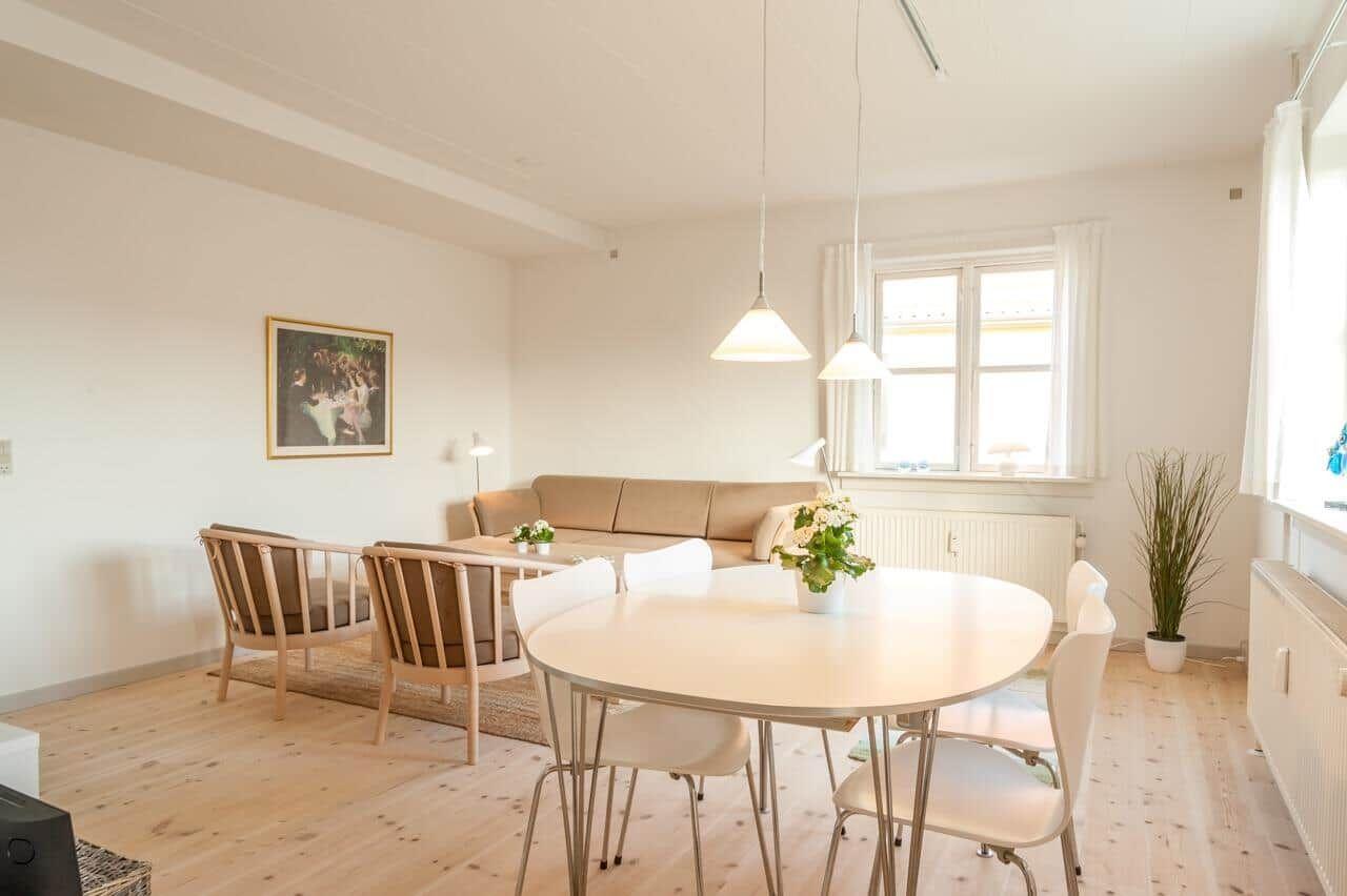 Ein Essbereich mit runden Tisch und weißen Stühlen. Im Hintergrund befindet sich ein Sofa und ein Fenster.