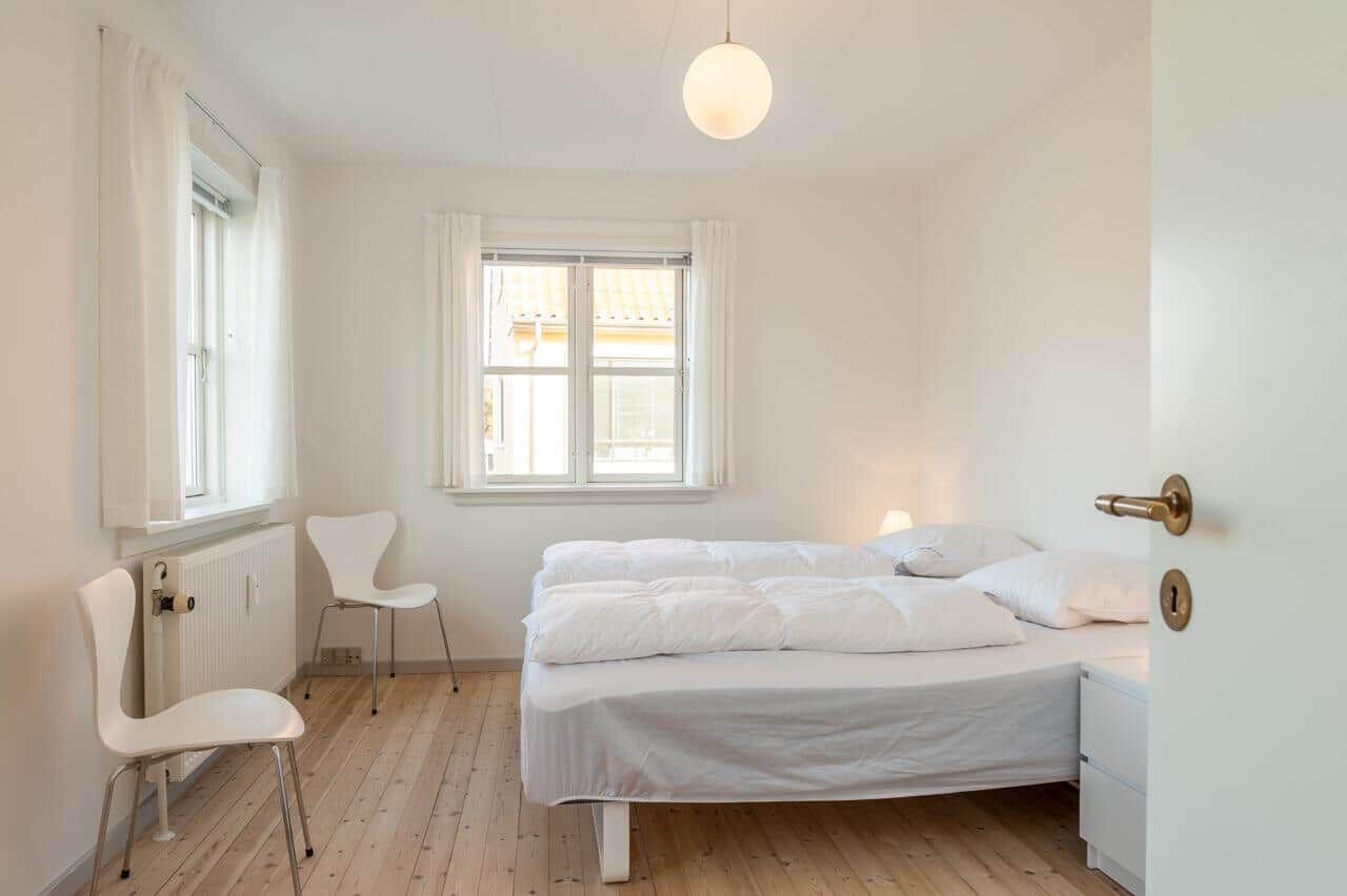 Schlafzimmer mit weißem Bett, zwei Fenstern und Holzboden.
