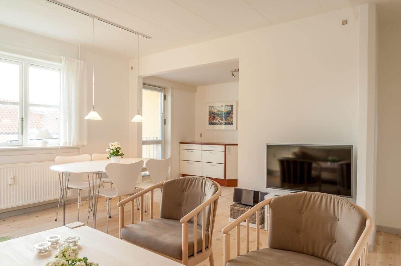 Wohn- und Essbereich mit TV, Stühlen und Tisch. Fenster mit Vorhängen. Holzboden.