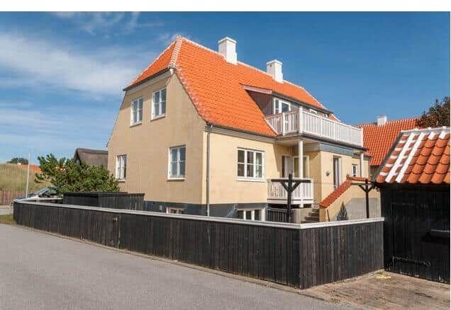 Haus mit rotem Dach, weißen Fenstern und Balkon. Umzäunt mit dunklem Holzzaun.