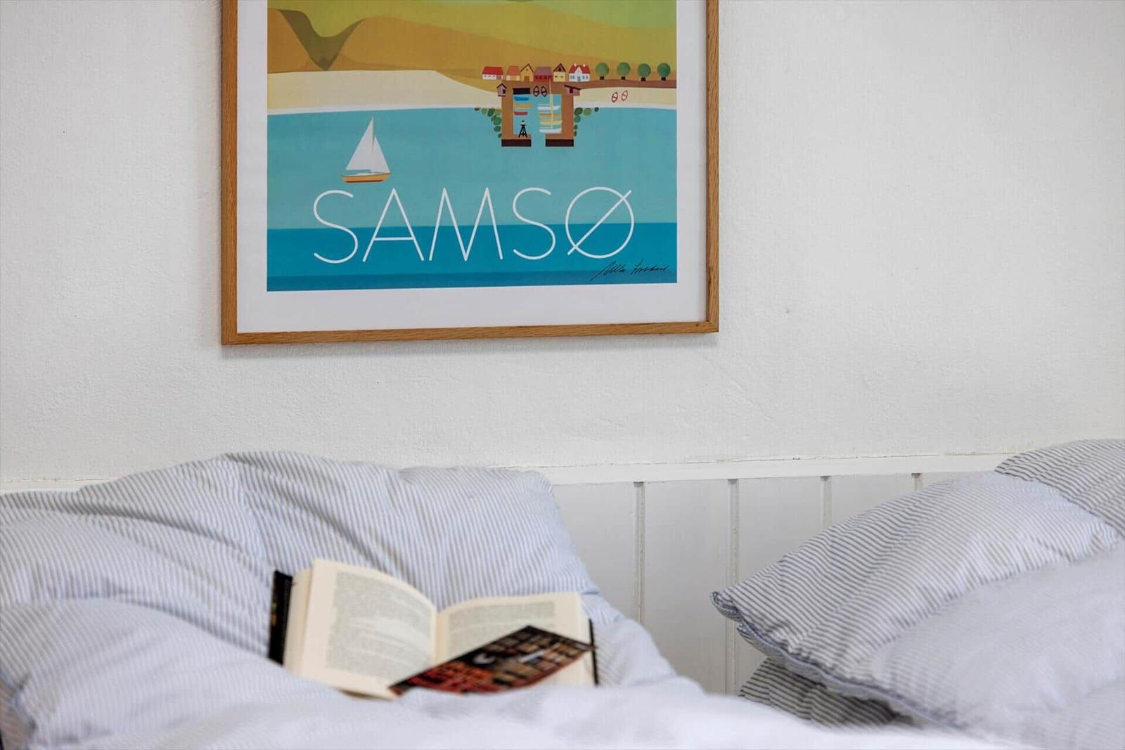 Ein Bett mit gestreiften Kissen und einem Buch. Über dem Bett hängt ein Bild mit der Aufschrift SAMSO.