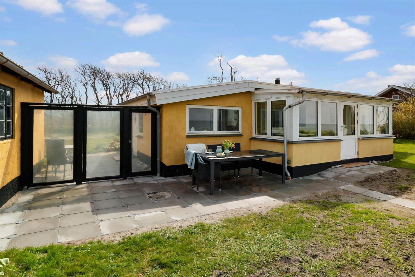 Gelbes Haus mit Terrasse, Tisch und Stühlen. Hinterhof mit Gras und Bäumen.