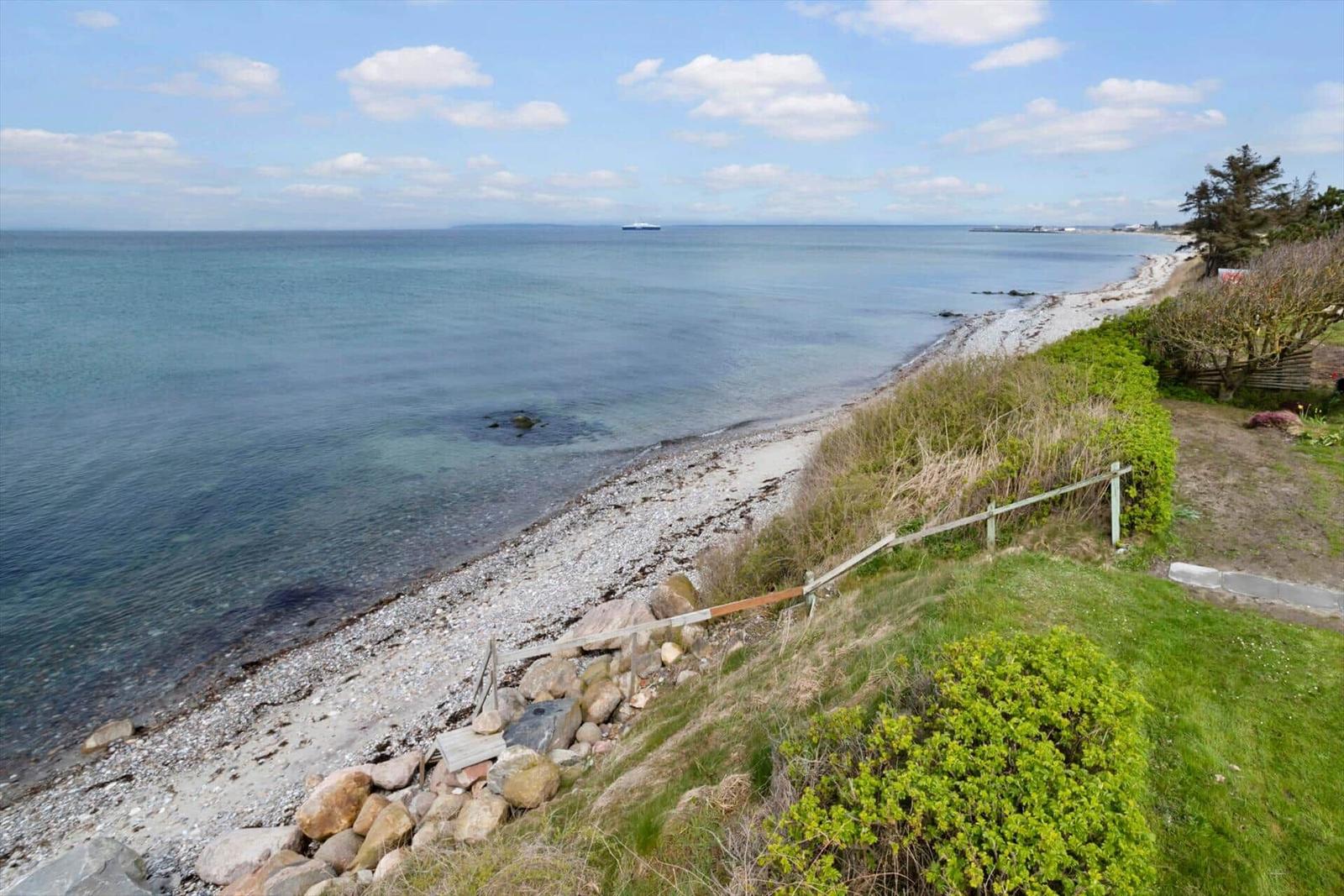Meerstrand mit Steinschotter und Grünflächen, Blick auf das Wasser und einen Schiff im Hintergrund.