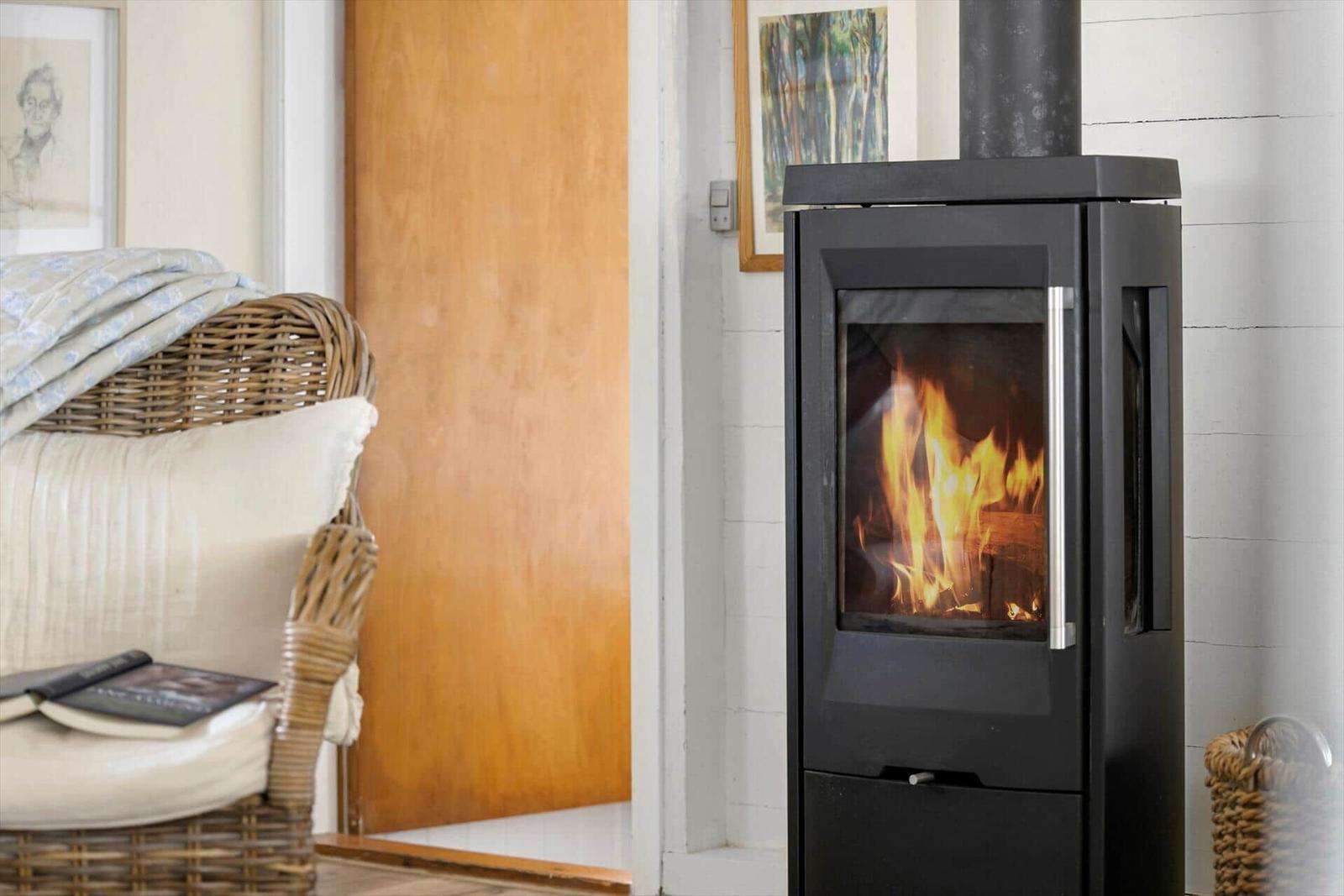 Ein Kaminofen mit brennendem Holz steht neben einem Strohhocker mit Kissen und Decke.