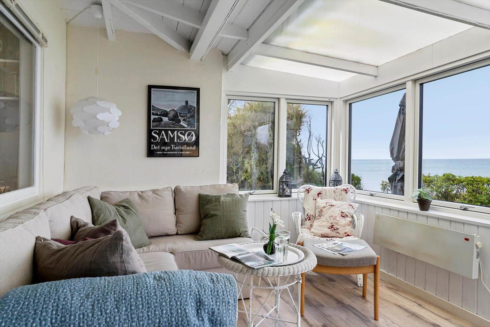Ein Wohnbereich mit Sofa, Sessel und Blick aufs Meer durch große Fenster.