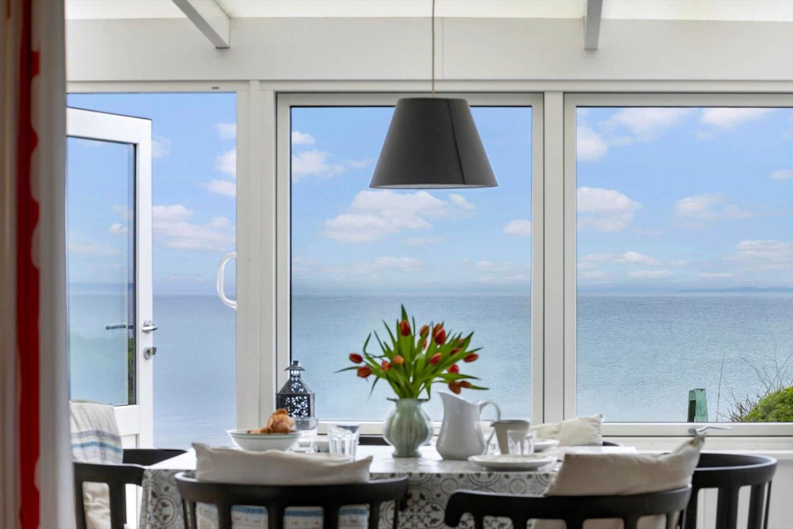 Esszimmer mit Meerblick durch große Fenster und Tisch mit Blumen.