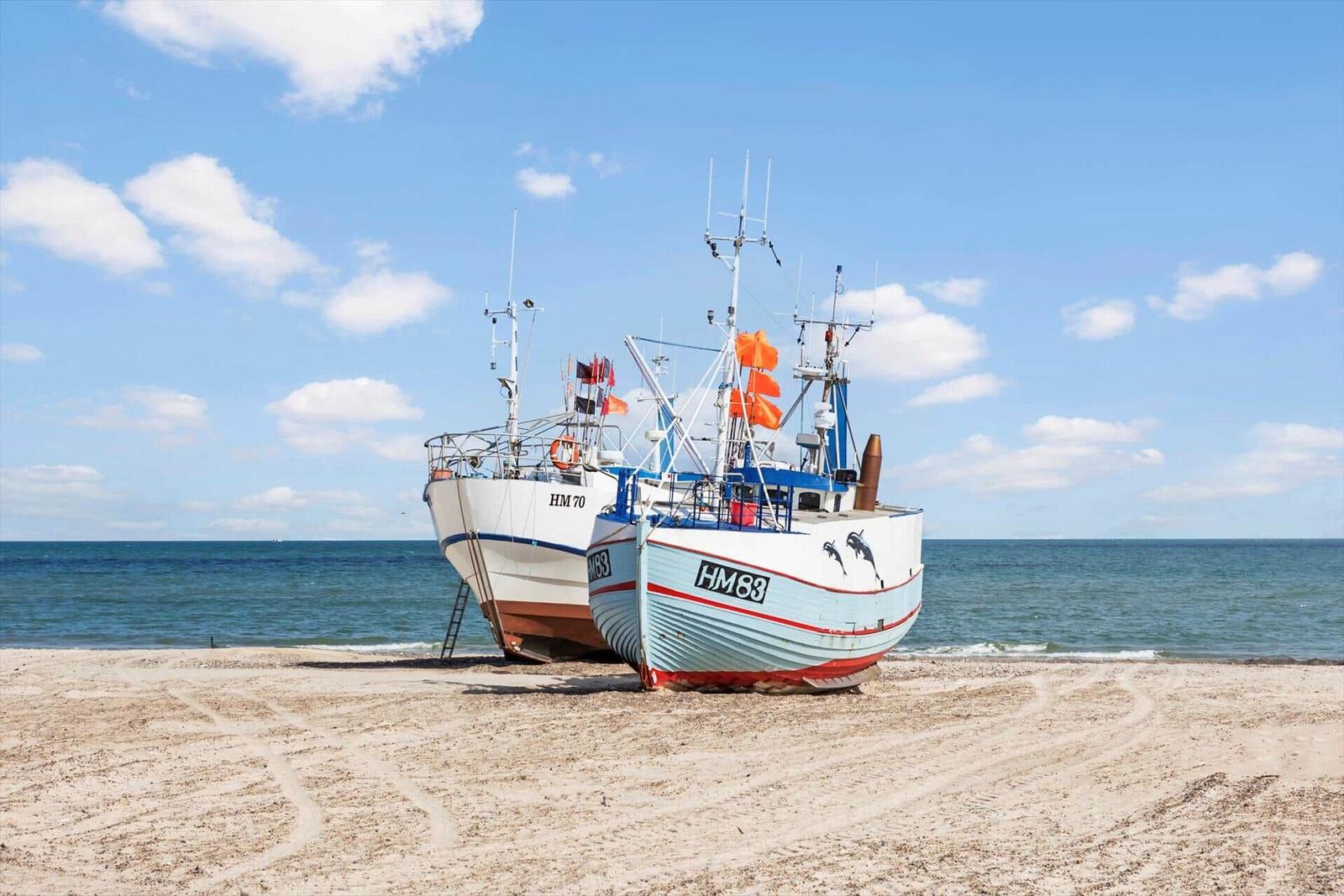 Zwei Fischerboote HM70 und HM83 liegen am Strand. Hinter ihnen erstreckt sich das Meer unter blauem Himmel.