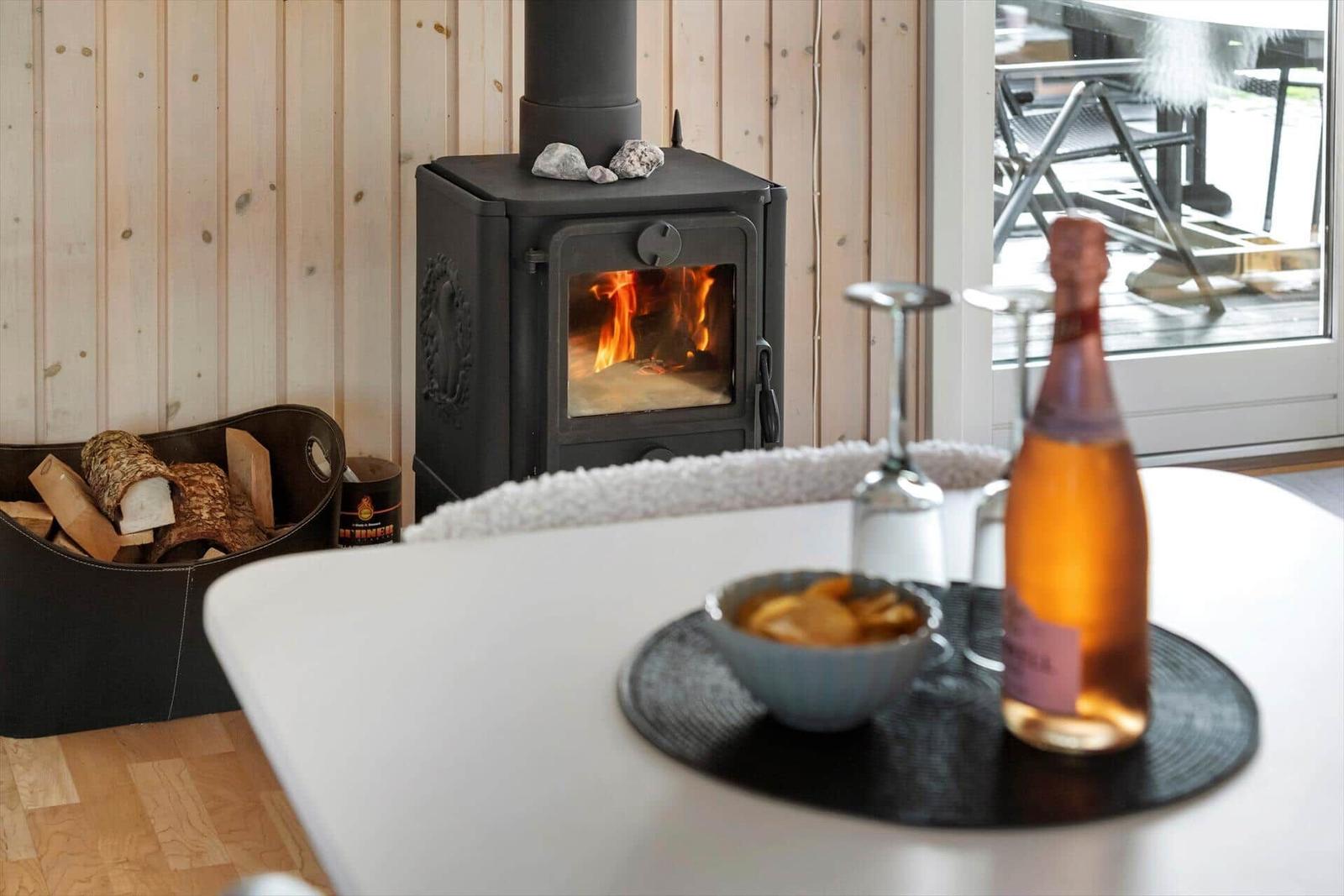 In the living area, a wood stove with a burning fire is visible. On the table, there is a glass, a bottle, and a bowl.