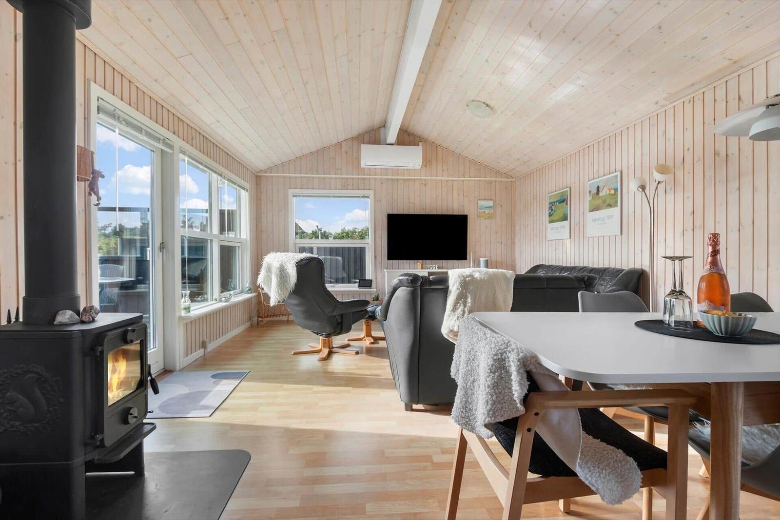 Living room with wooden ceiling, wood stove, sofa, and dining table. Large windows and TV visible.
