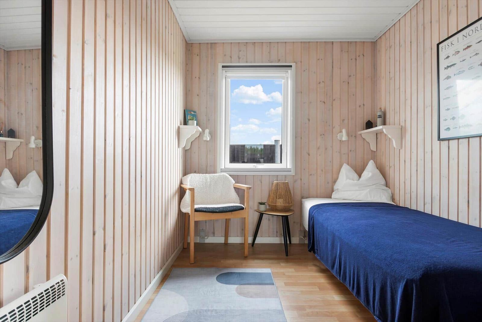 A bedroom with wooden walls, bed with blue bedding, chair, and window.