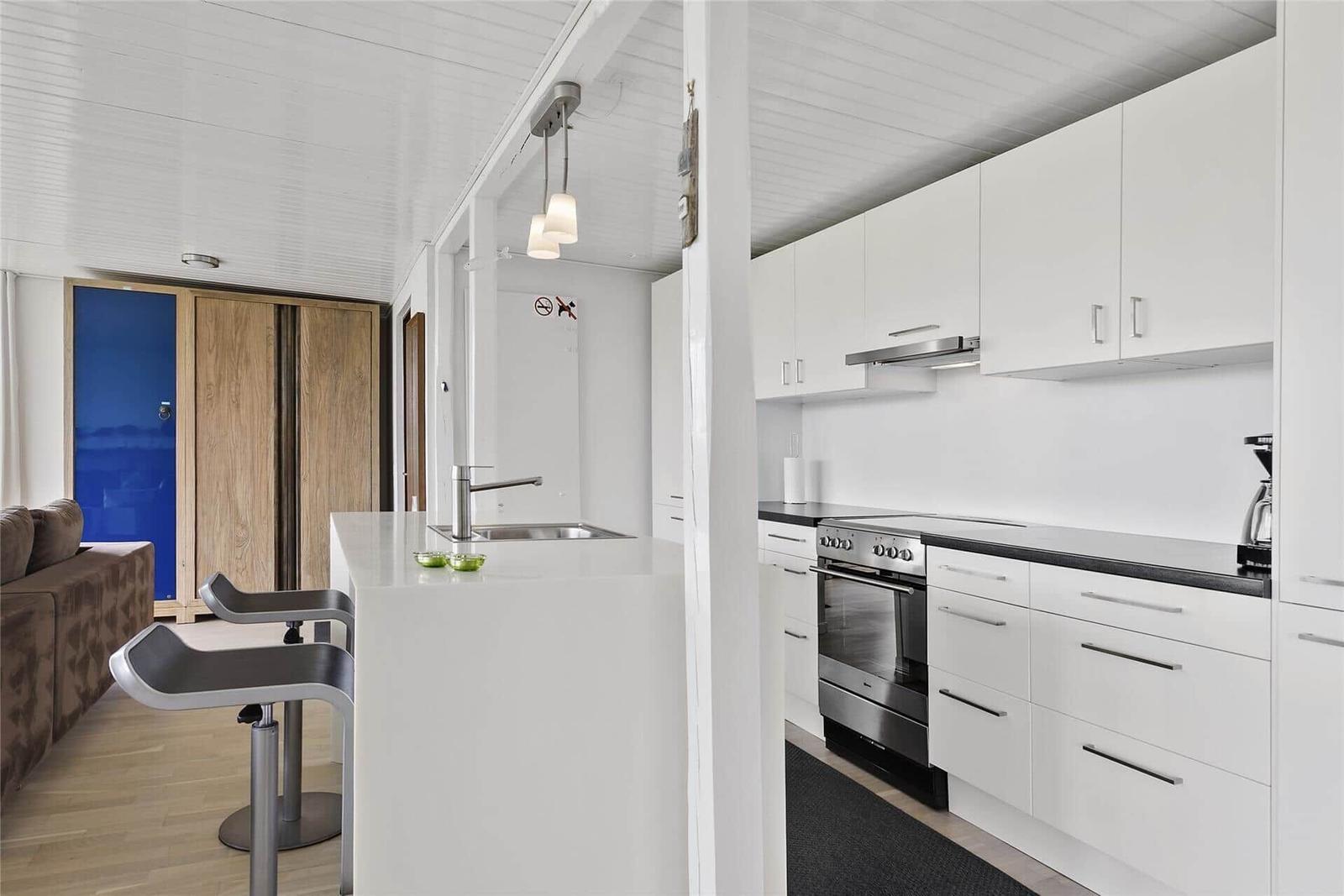 Küche mit Arbeitsplatte, Spüle und Kochfeld. Im Hintergrund Wohnbereich mit Sofa und Tür.