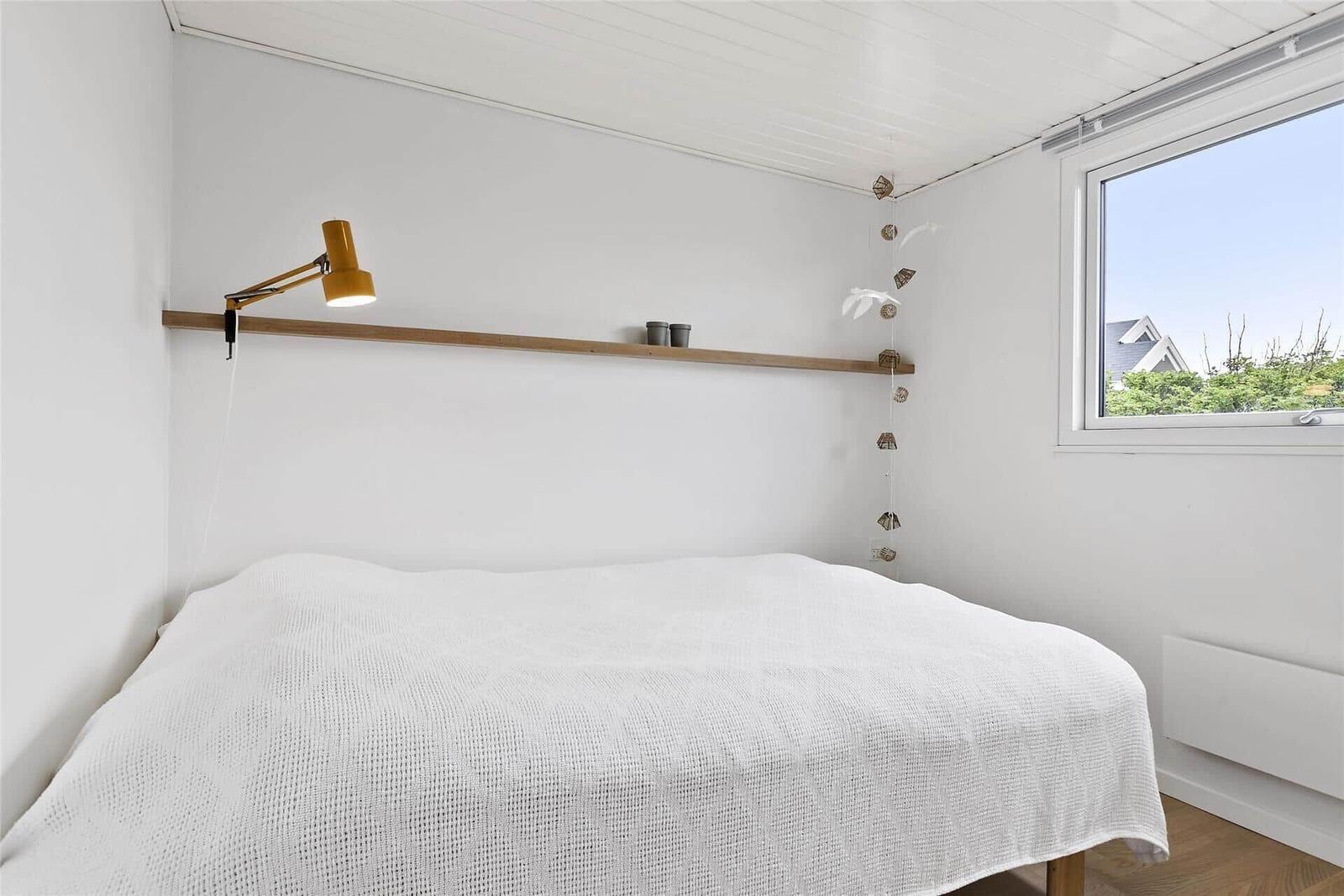 Schlafzimmer mit weißem Bett, Holzregal und Fenster. Decke mit Holzverkleidung.