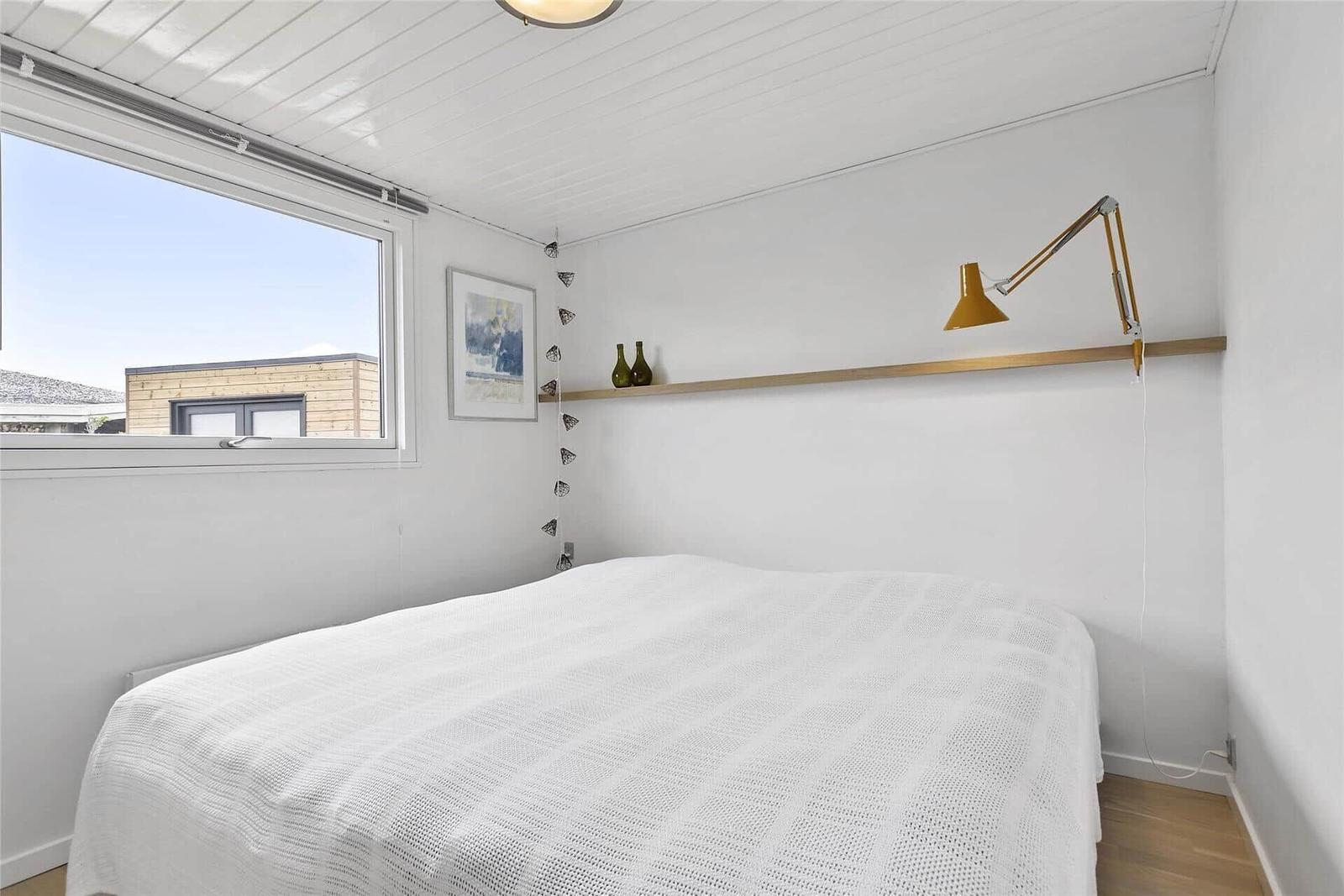 Schlafzimmer mit weißem Bett, Fenster und Wandlampe.