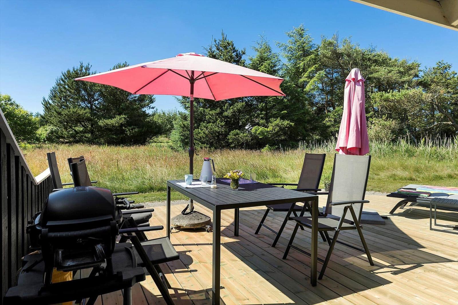Terrasse mit Tisch, Stühlen, Sonnenschirm und Grill. Hintergrund: Gras und Bäume.