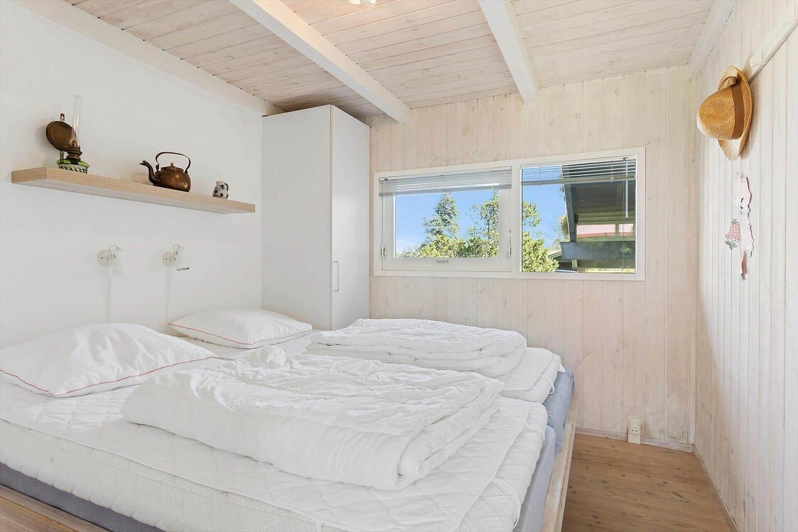 Schlafzimmer mit Bett, Fenster und Holzwand. Auf Regal stehen Teekanne und Laterne.