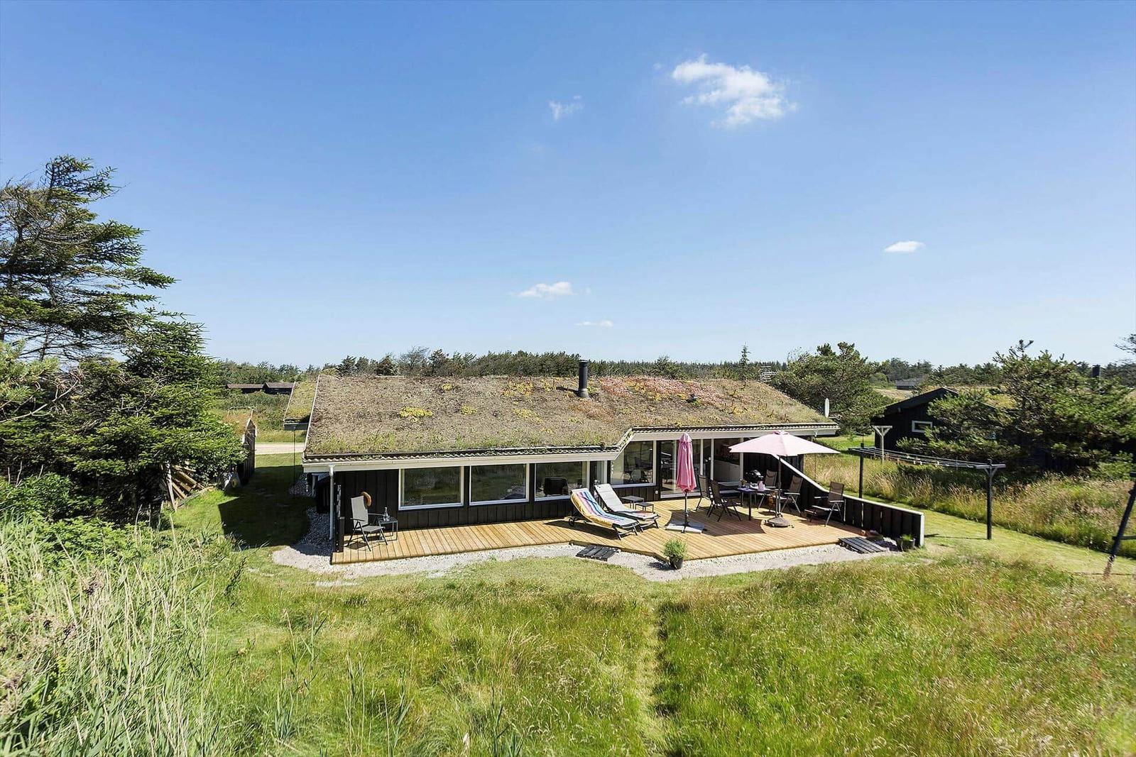 Haus mit grüner Dachdecke, Terrasse mit Sonnenliegen und Tisch, umgeben von Gras und Bäumen.
