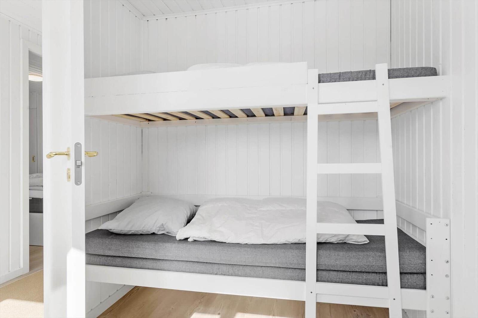 Ein Doppelstockbett mit grauen Matratzen und weißen Kissen in einem Raum mit weiß verkleideten Wänden.