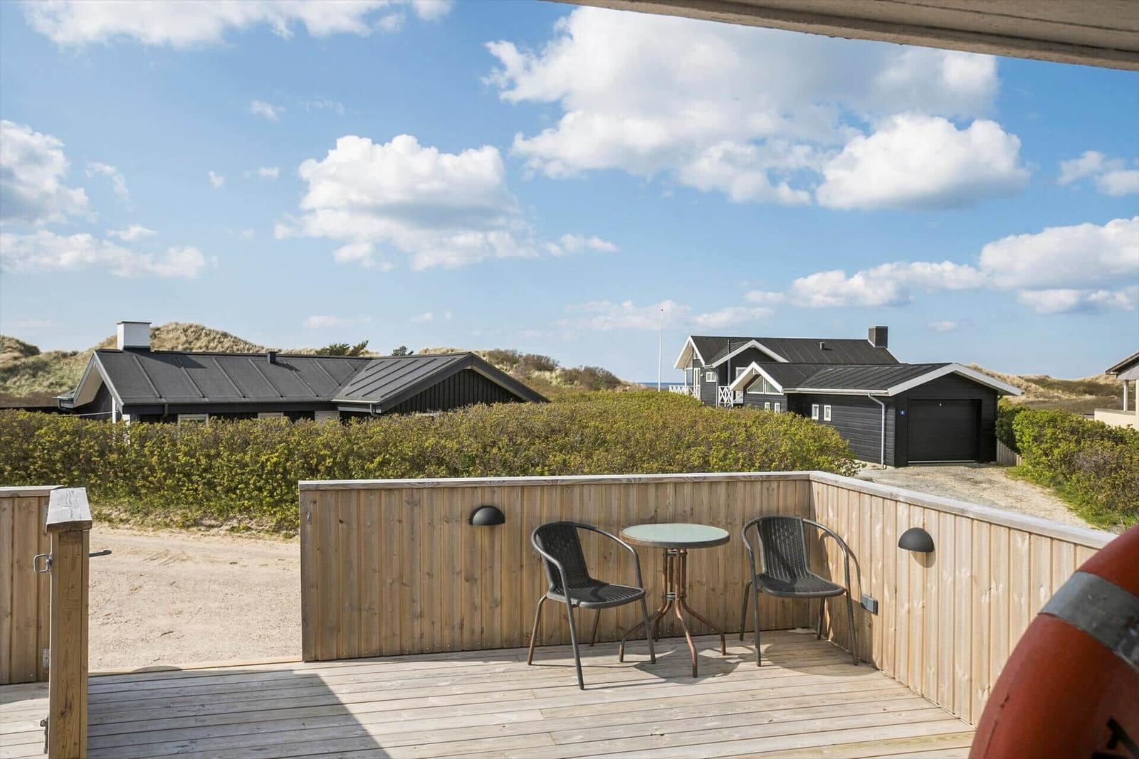 Terrasse mit Tisch und zwei Stühlen. Blick auf Häuser und Dünen unter blauem Himmel.