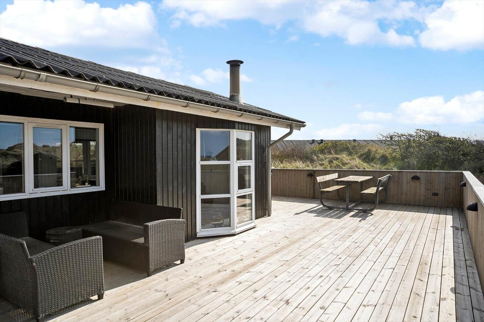 Terrasse mit Holzdielen, Sitzgruppe und Tisch. Dunkel verkleidetes Haus mit Fenstern und Schornstein.