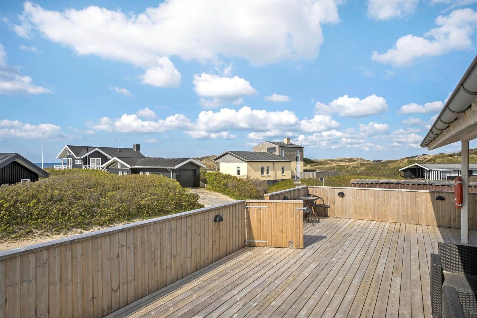 Holzterrasse mit Blick auf Strandhäuser und Dünen unter blauem Himmel.
