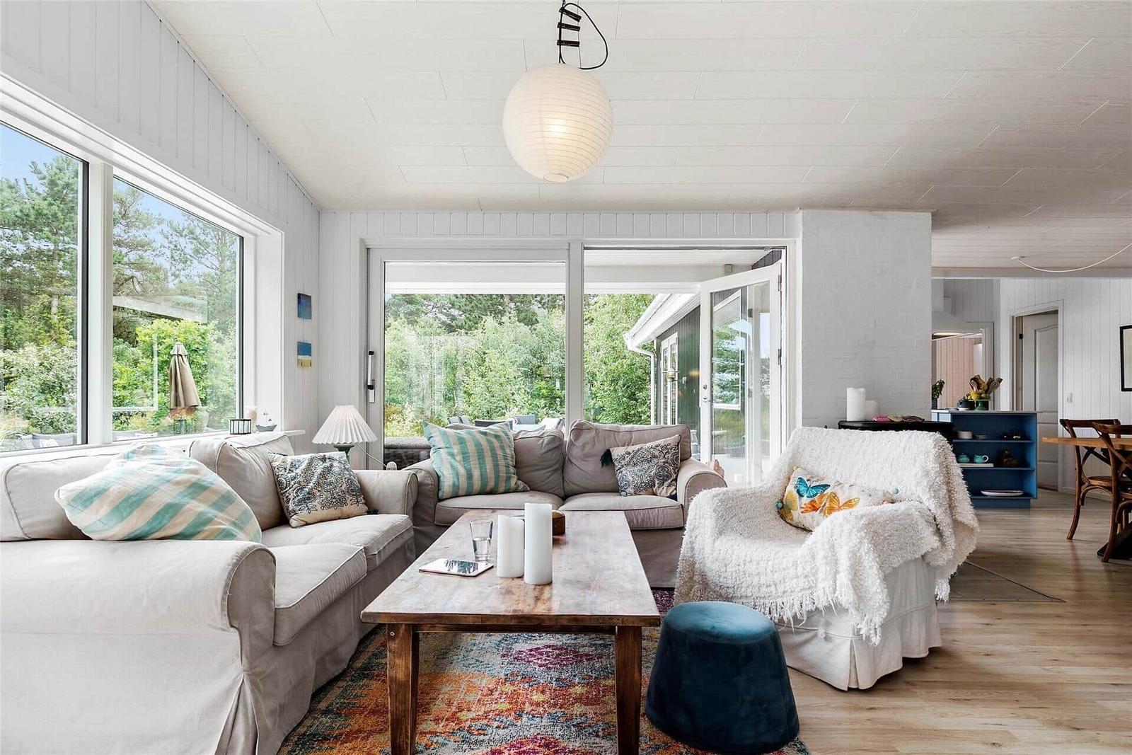 Wohnzimmer mit Sofa, Sessel und Holztisch. Große Fenster und Glastür zur Außenterrasse.
