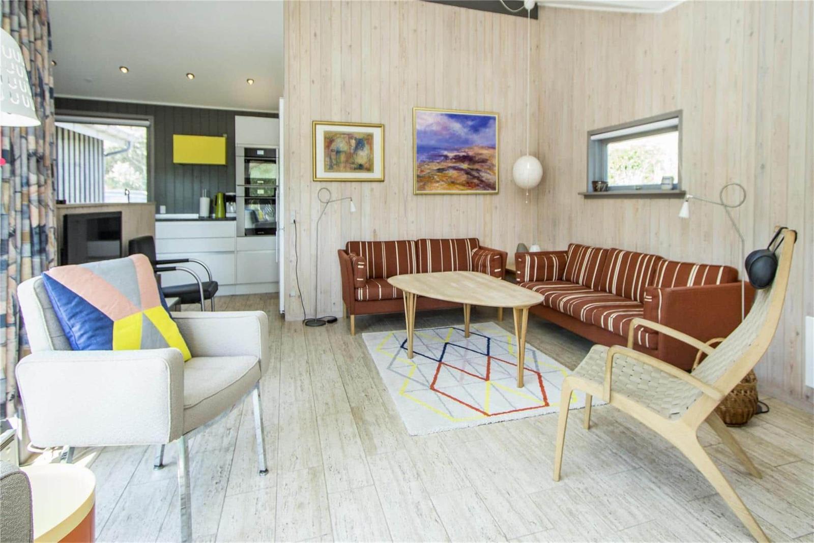 Wohnbereich mit Holzwänden, Sofa, Tisch und Sessel. Küche im Hintergrund sichtbar.