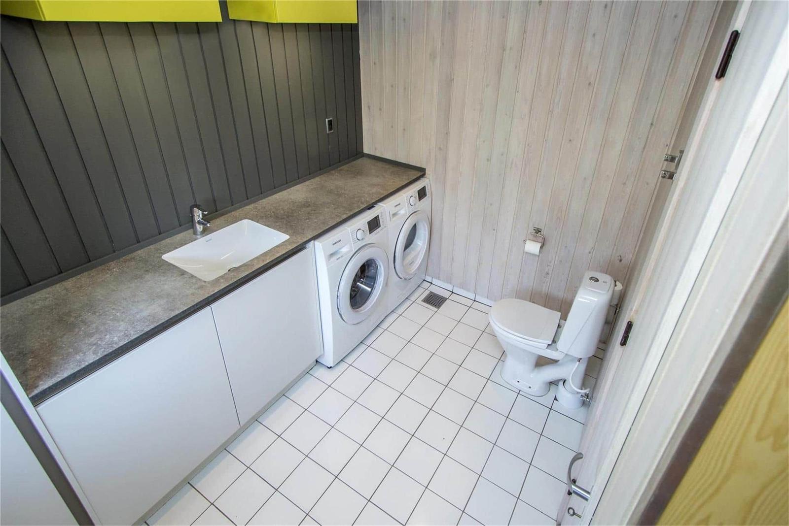 Waschraum mit Waschmaschine, Spülung und Toilette. Holzwand und Fliesenboden.