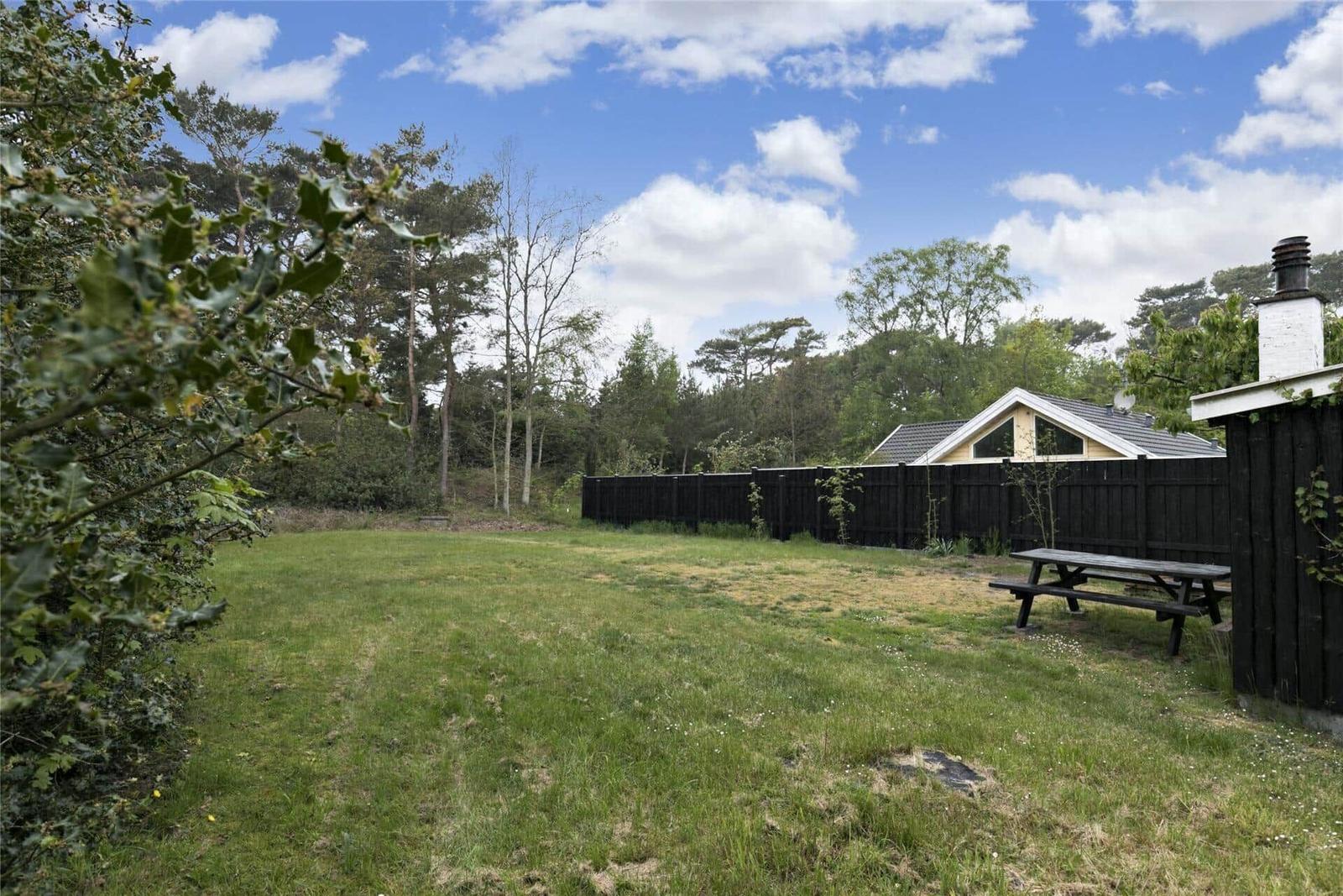 Grüner Garten mit Picknicktisch, umgeben von Wald und Zaun. Ferienhaus im Hintergrund.