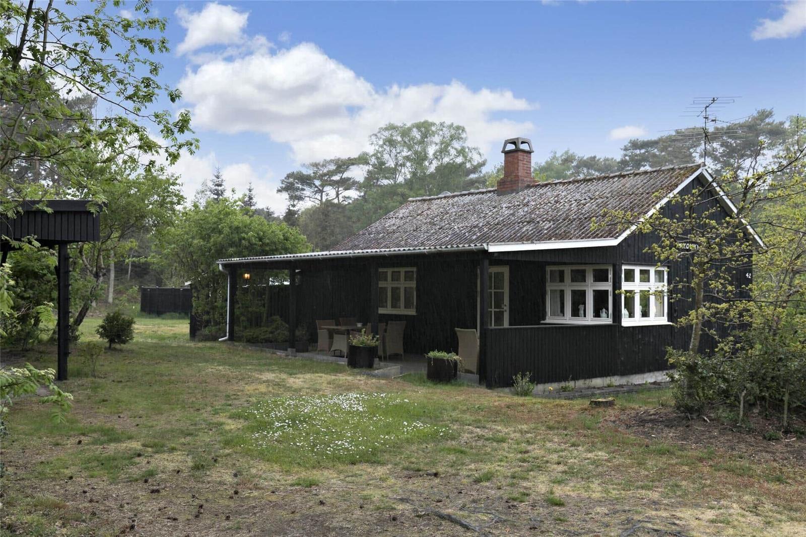 Schwarzes Holzhaus mit Terrasse und Garten. Fenster mit weißen Rahmen. Hintergrund Wald.