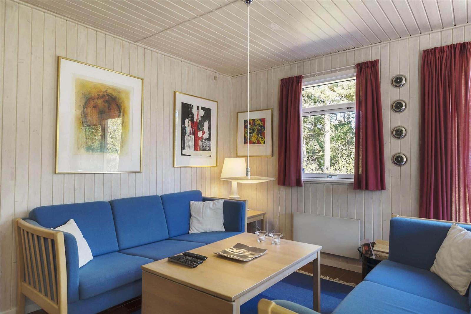 Ein Wohnzimmer mit blauen Sofas, Holztisch und Wandbildern. Rote Vorhänge und Fensterblick.