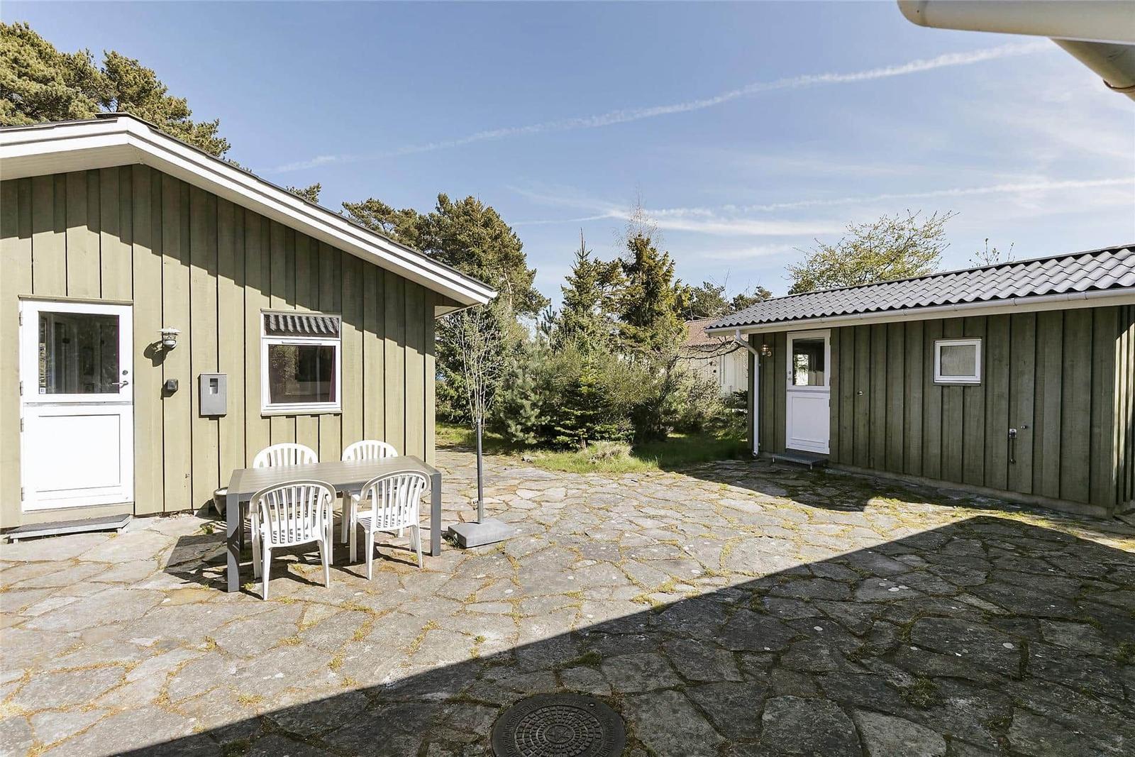Haus mit Steinplattenhof, weiße Tische und Stühle, umgeben von Bäumen.