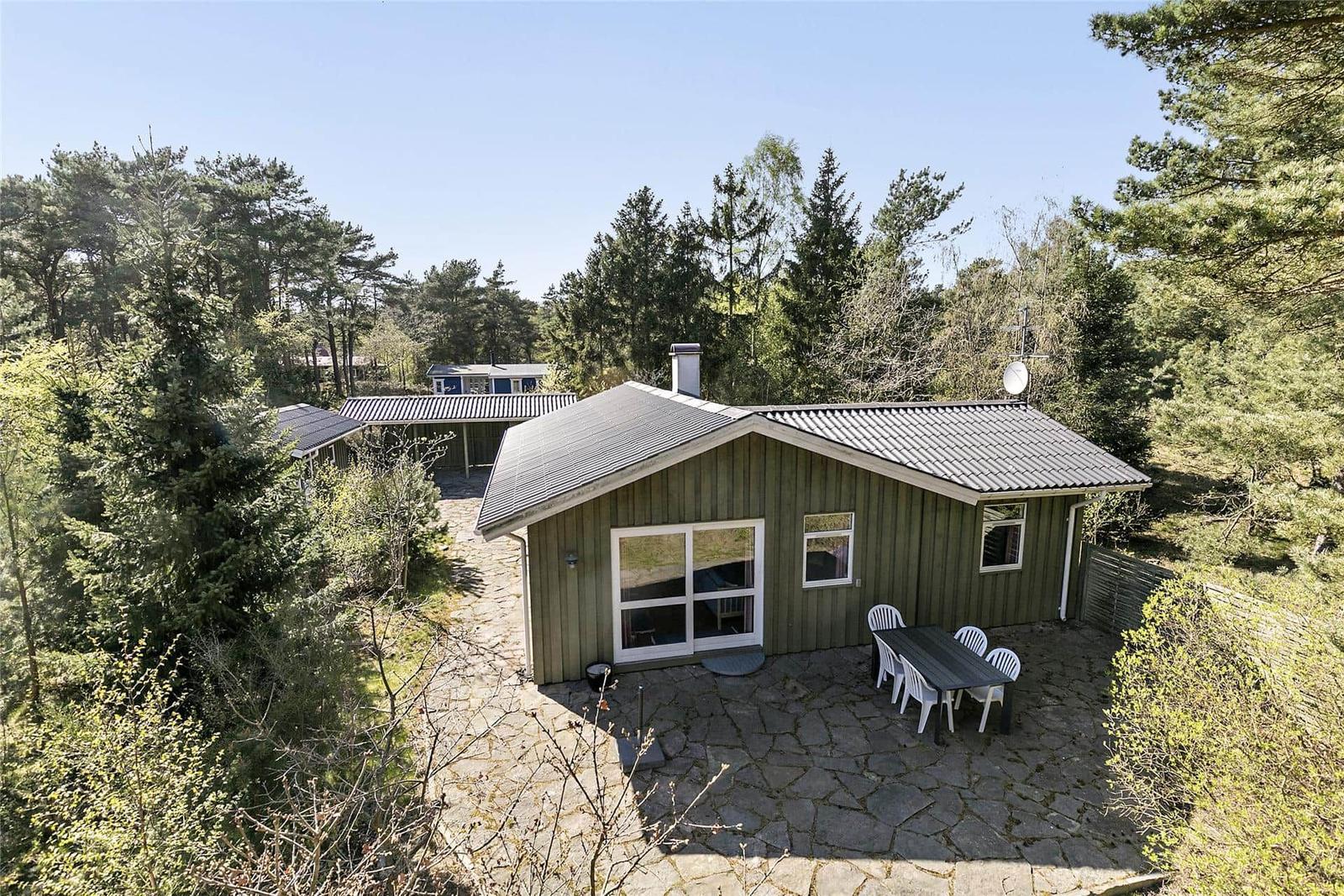 Grünes Holzhaus mit Terrasse und Garten. Umgeben von Bäumen. Tisch und Stühle draußen.