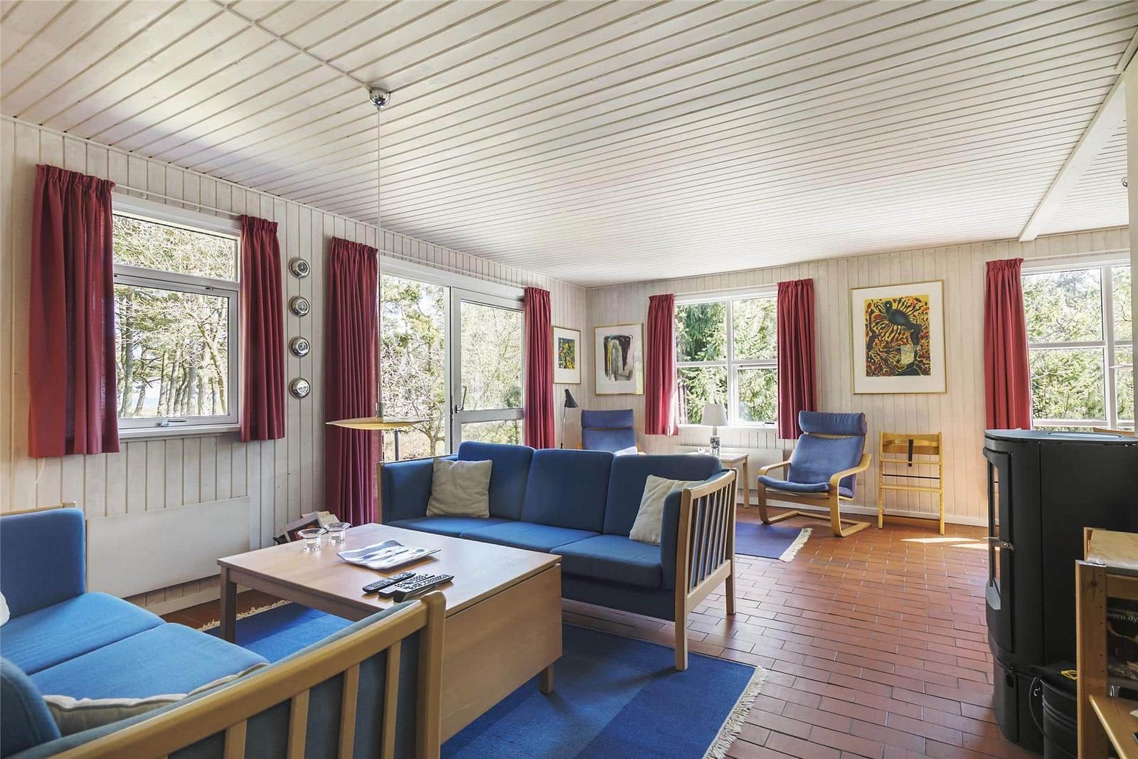 Wohnzimmer mit blauem Sofa, Holzmöbeln und roten Vorhängen an großen Fenstern.